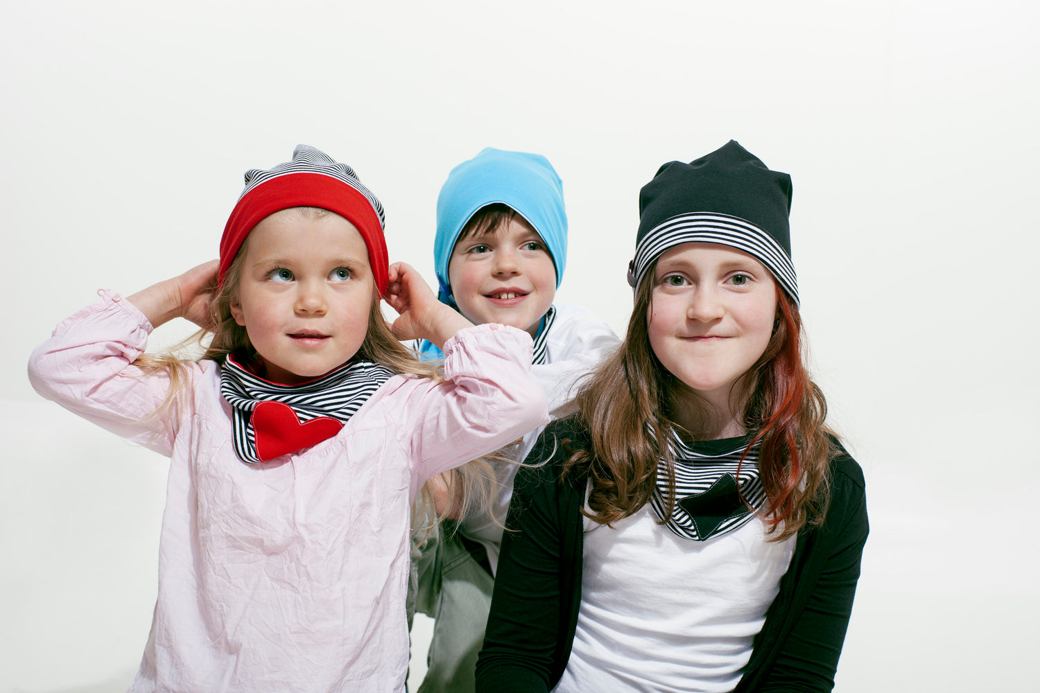 3 Kinder, 2 Mädchen und ein Junge in ihrer Mitte sitzen auf dem Boden. Sie tragen geringelte Mützen mit passenden Halstüchern.