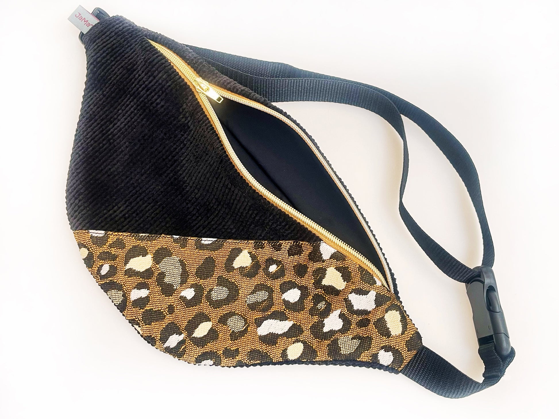 Eine Bauchtasche in abgerundeter Form. Das Vorderteil ist geteilt. Der linke Teil ist aus schwarzem Breitcord und der rechte aus einem Jacquard Canvas im Leopradenprint. Die Tasche wird mit einem goldenen Reißverschluss auf braunem Band geschlossen. Der Deckel der Tasche ist aus dem schwarzen Breitcord. Sie hat schwarzes Gurtband, eine Steckschnalle und einen Versteller aus schwarzem Kunststoff. 
Der Reißverschluss ist ein Stückk geöffnet, so dass das schwarze Futter zu sehen ist.
Ansicht von oben, schräg.