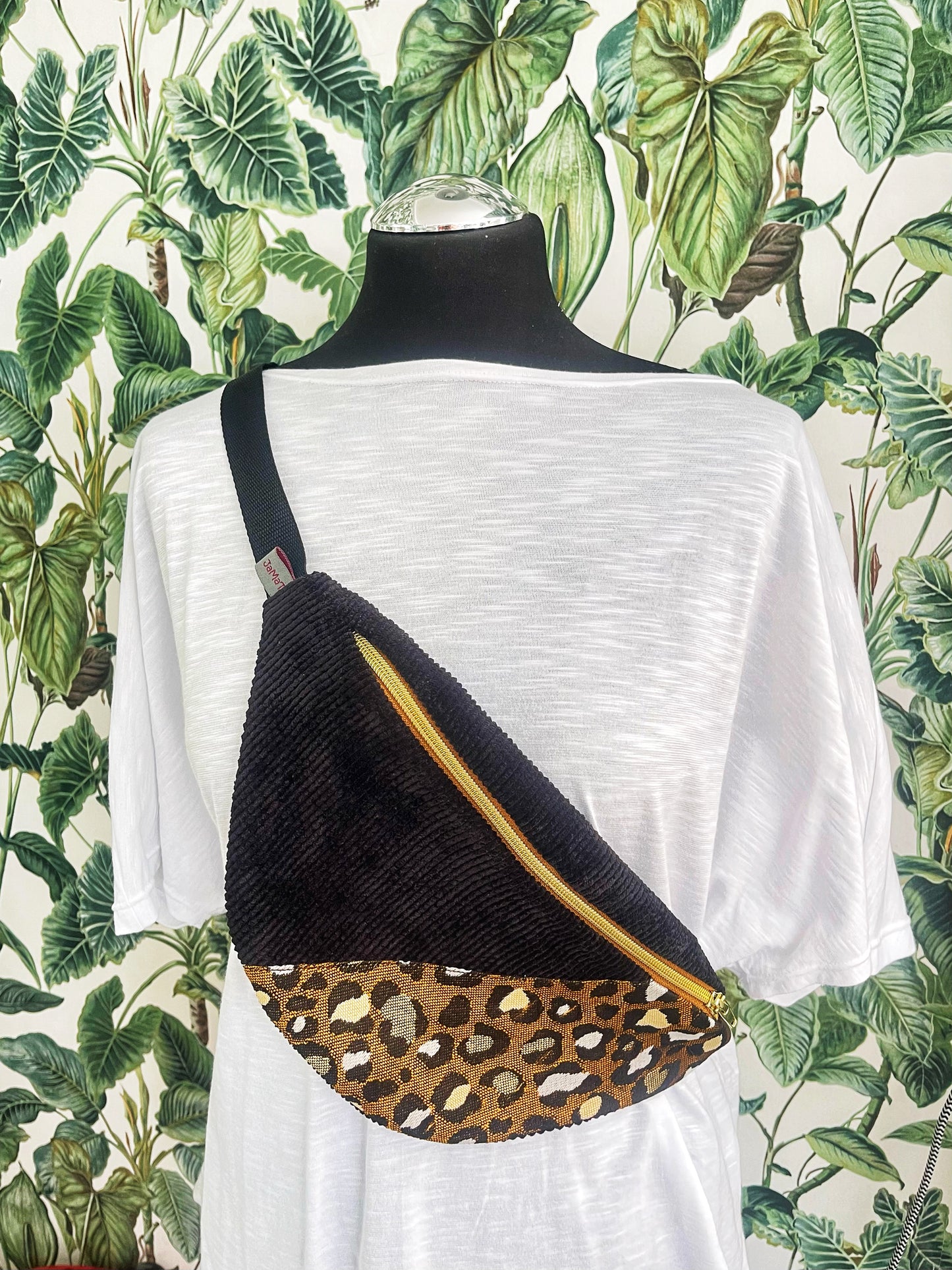 Die Bauchtasche  angezogen auf einer Schneiderpuppe, die ein weißes T-Shirt trägt. Die Schneiderpuppe selbst ist schwarz und steht vor einer Tapete mit grünen Blättern. Sie hat die Bauchtasche schräg nach vorne umgehängt.