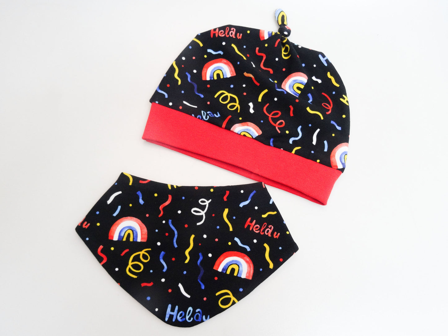 Set aus Zipfelmütze und Halstuch für Babys und kleine Kinder, schwarz mit buntem Schriftzug Helau, Konfetti, Luftschlangen und Regenbögen. Ansicht liegend von oben.