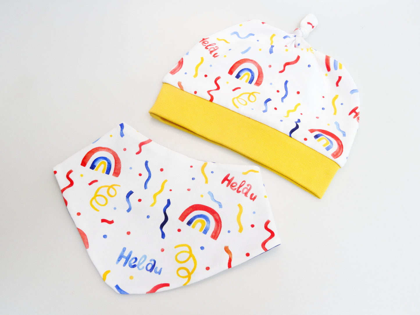 Set aus Zipfelmütze und Halstuch für Babys und kleine Kinder weiß mit buntem Schriftzug Helau, Konfetti, Luftschlangen und Regenbögen. Ansicht liegend von oben.