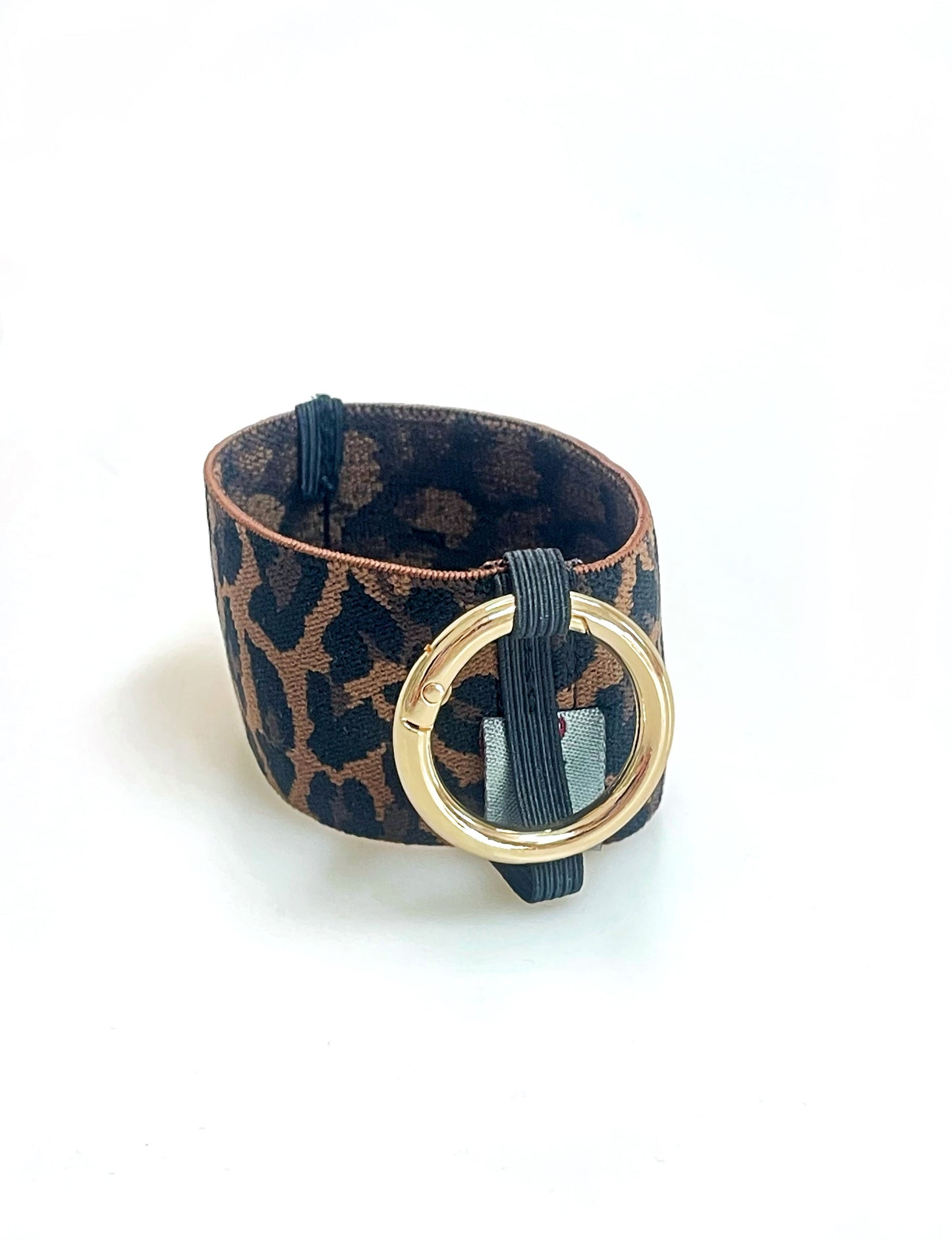 Ein Becherhalter aus einem breiten Gummiband mit Leoparden Muster. Er hat einem runden, goldenen Karabinerhaken. Das Gummiband ist zu einem Kreis zusammengenäht. Der Karabiner ist an einem schmalen Gummiband befestigt, das unten um das breite Gummiband herum geht.