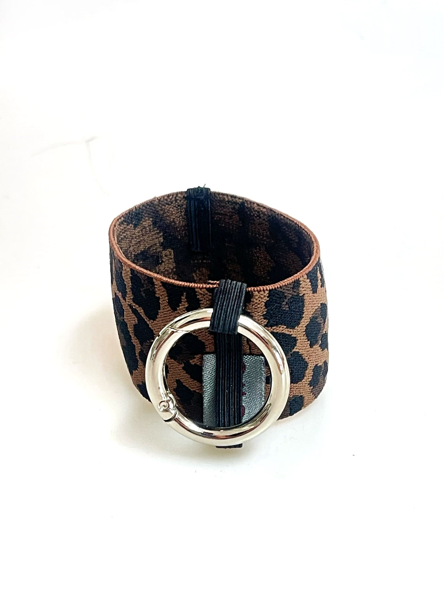Ein Becherhalter aus einem breiten Gummiband mit Leoparden Muster. Er hat einem runden, silbernen Karabinerhaken. Das Gummiband ist zu einem Kreis zusammengenäht. Der Karabiner ist an einem schmalen Gummiband befestigt, das unten um das breite Gummiband herum geht.