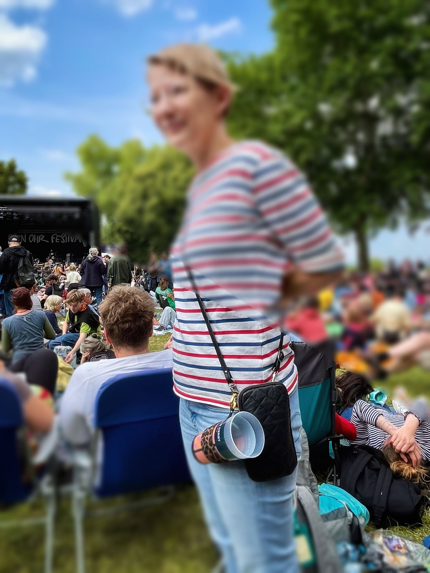 Der Becherhalter mit Leopardenmuster ist an einer kleinen, schwarzen Tasche befestigt, die eine Frau in Jeans und Ringelshirt trägt. Es steckt ein leerer Pfandbecher darin. Unscharf sind andere Leute , Bäume, eine Festivalbühne und blauer Himmel zu erkennen.