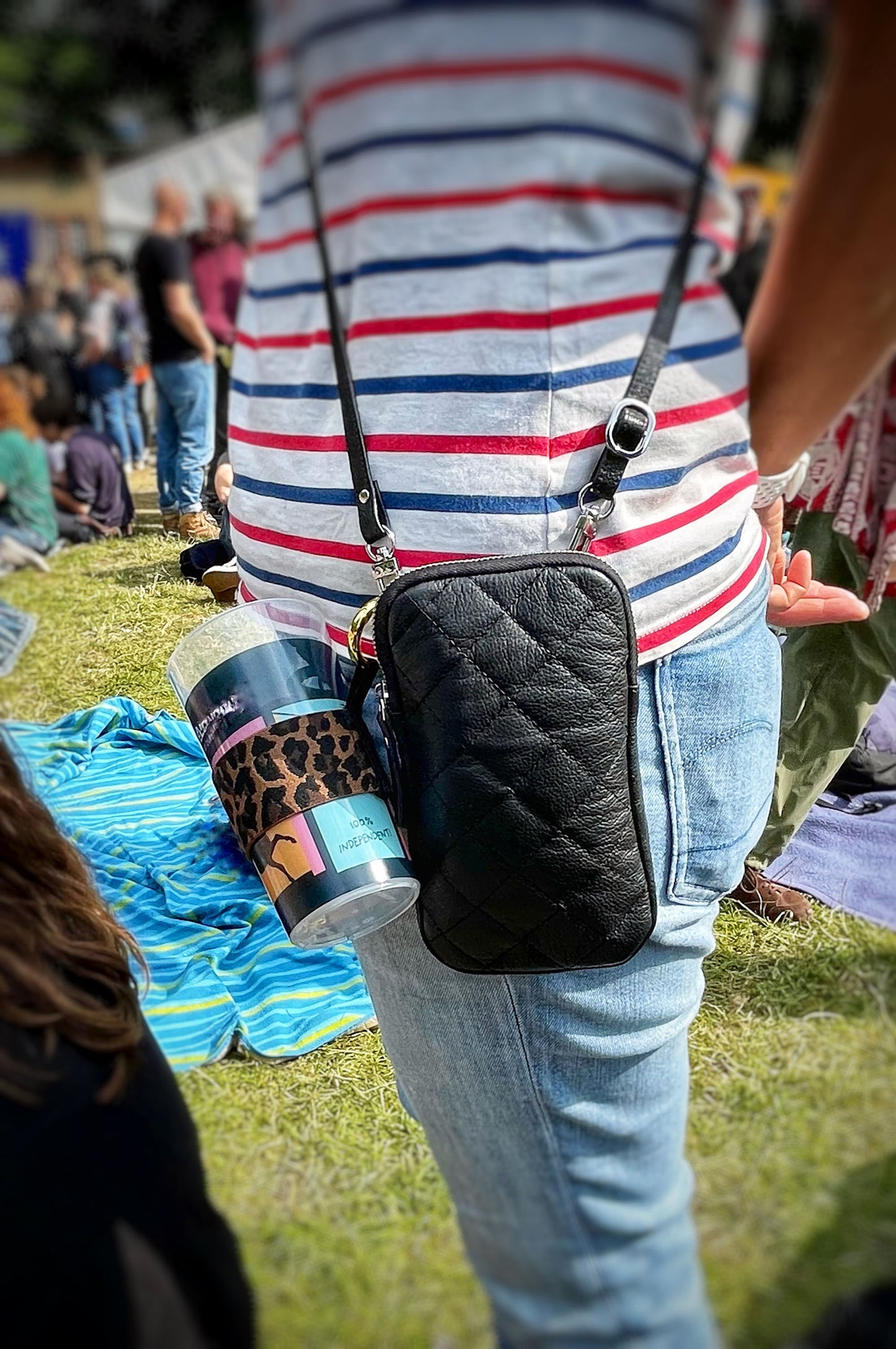 Der Becherhalter mit Leopardenmuster ist an einer kleinen, schwarzen Tasche befestigt, die eine Frau iin Jeans und Ringelshirt trägt. Es steckt ein leerer Pfandbecher darin. Unscharf sind andere Leute und rasen zu erkennen.