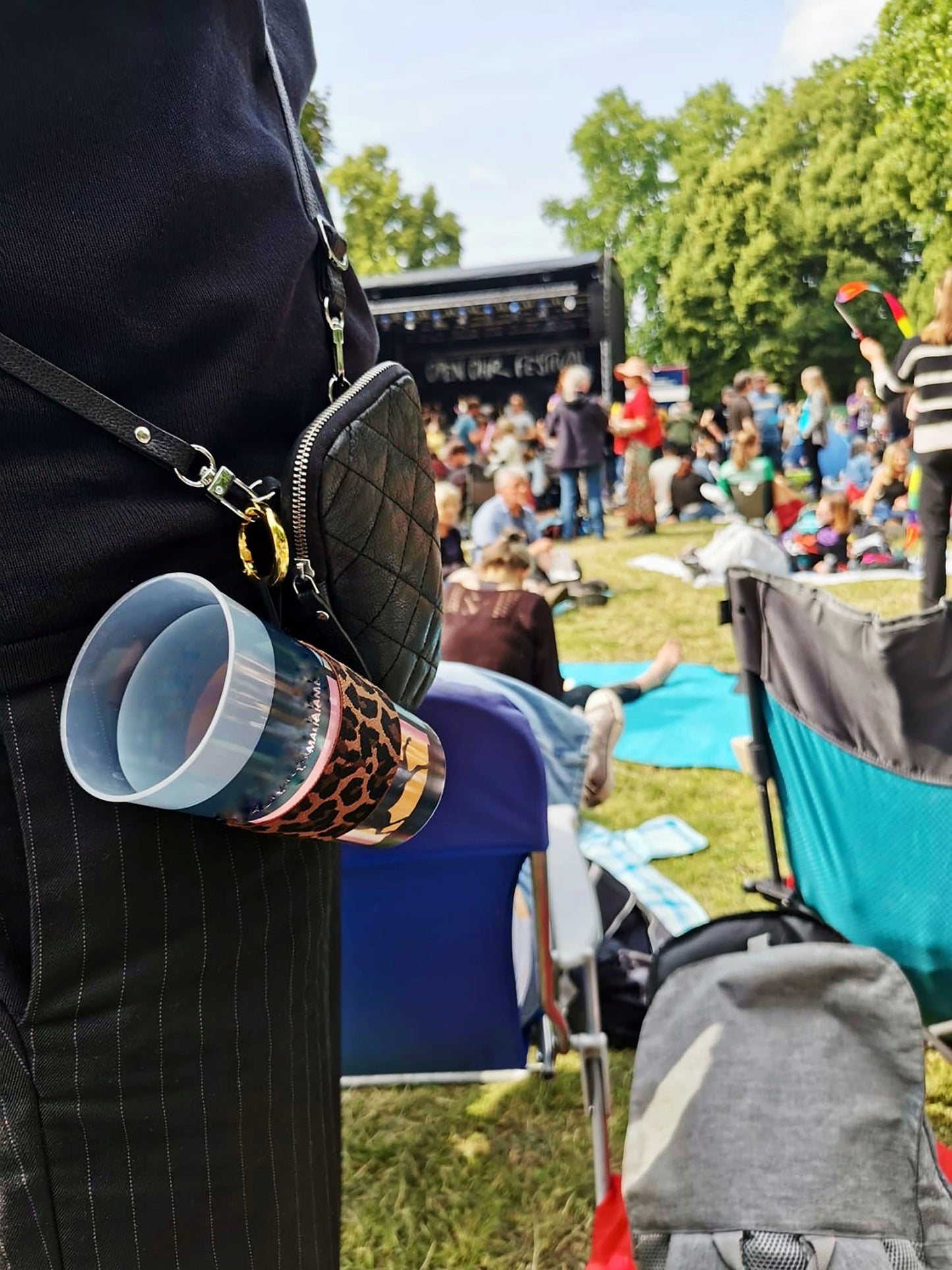 Der Becherhalter mit Leopardenmuster ist an einer kleinen, schwarzen Tasche befestigt, die eine Frau in schwarz trägt. Es steckt ein leerer Pfandbecher darin. Unscharf sind andere Leute , Bäume, eine Festivalbühne und blauer Himmel zu erkennen.