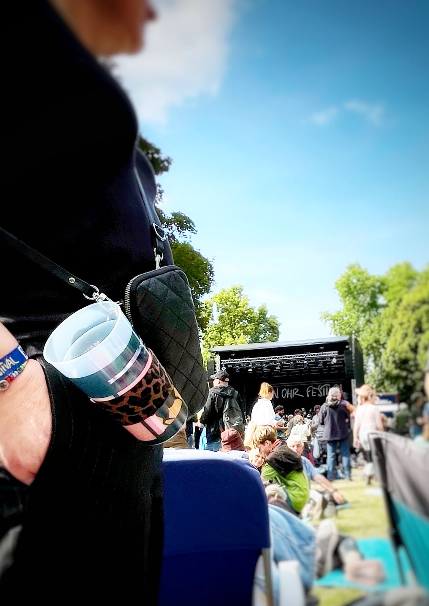 Der Becherhalter mit Leopardenmuster ist an einer kleinen, schwarzen Tasche befestigt, die eine Frau in schwarz trägt. Es steckt ein leerer Pfandbecher darin. Unscharf sind andere Leute , Bäume, eine Festivalbühne und blauer Himmel zu erkennen.
