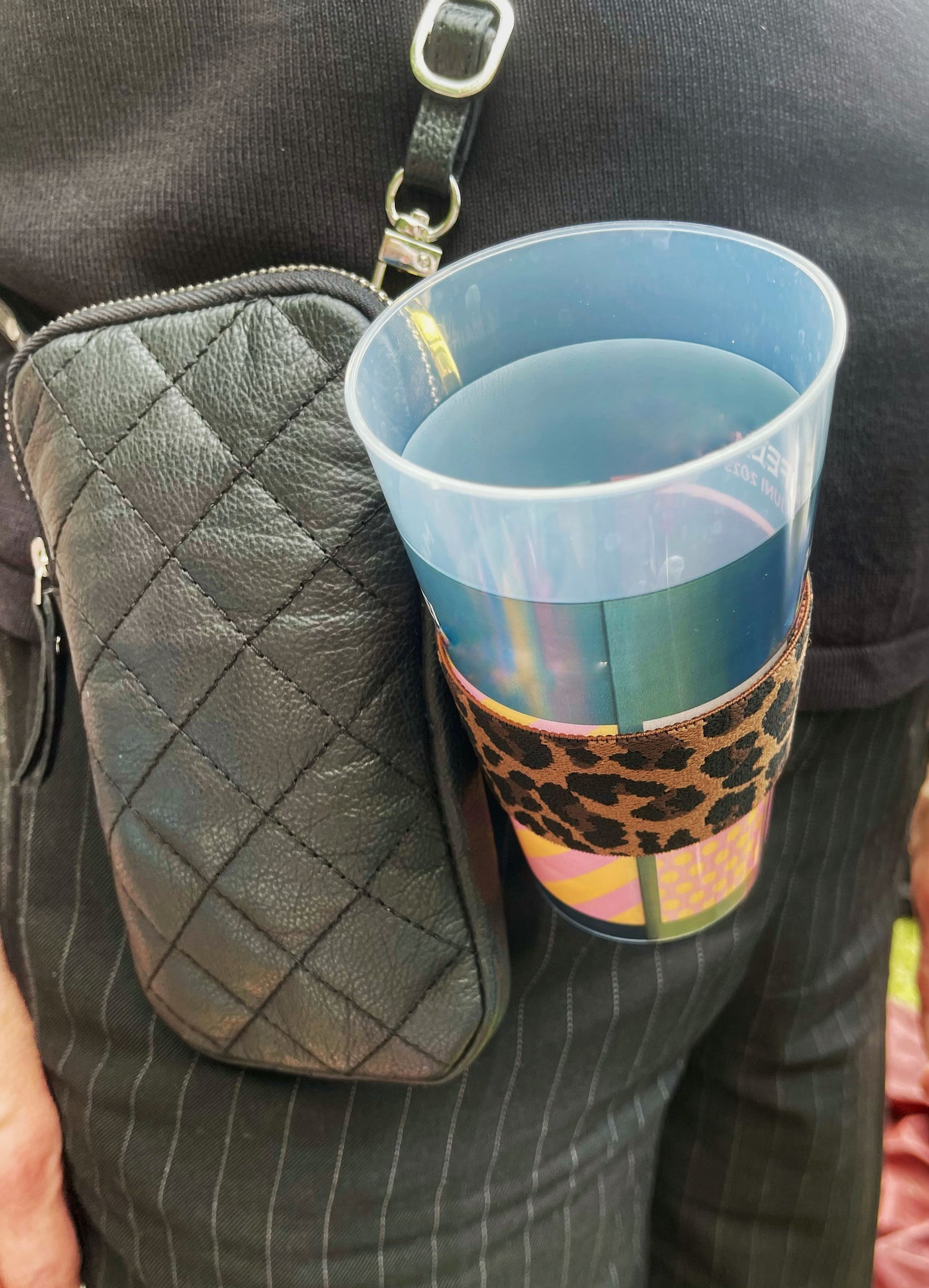 Der Becherhalter mit Leopardenmuster ist an einer kleinen, schwarzen Tasche befestigt, die eine Frau in schwarz trägt. Es steckt ein Pfandbecher aus Plastik darin. Nahaufnahme des Becherhalters mit Becher und Tasche.