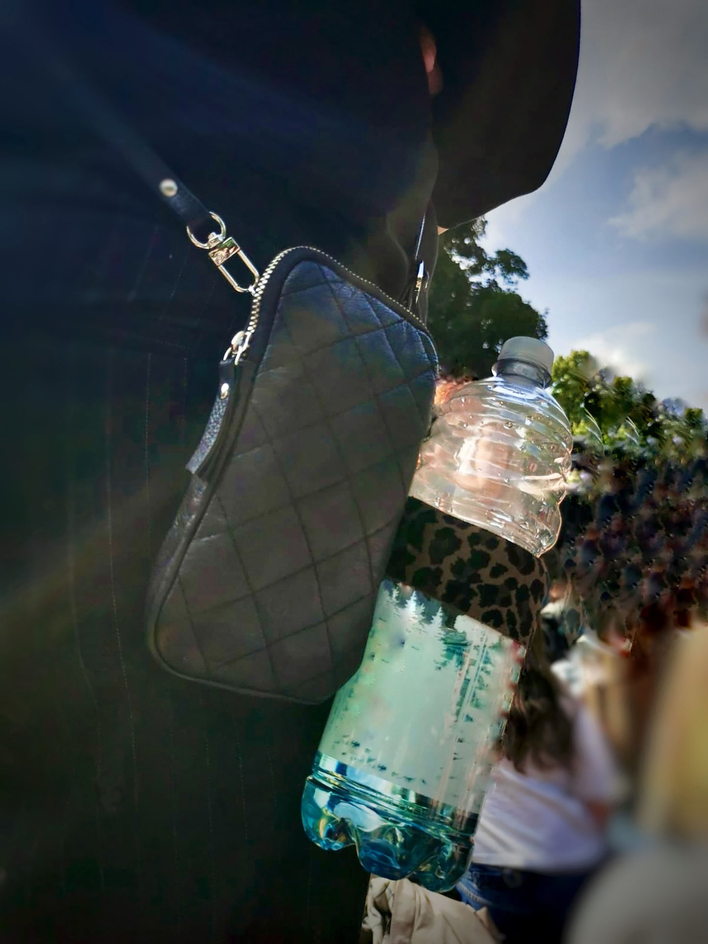 Der Becherhalter mit Leopardenmuster ist an einer kleinen, schwarzen Tasche befestigt, die eine Frau in schwarz trägt. Es steckt eine 1 Liter Wasserfalsche darin. Unscharf sind andere Leute , Bäume und blauer Himmel zu erkennen.