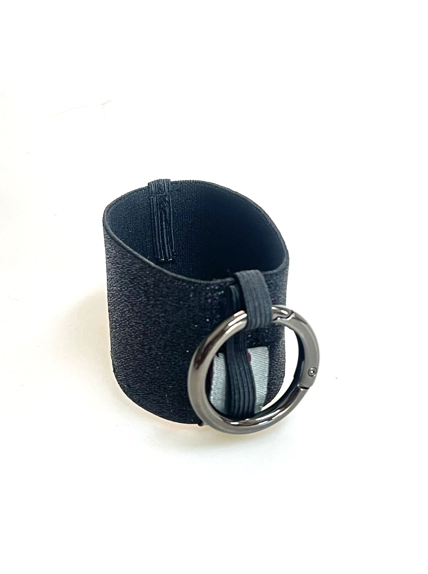 Ein Becherhalter aus einem gold glitzernden, breiten Gummiband, innen schwarz, mit einem runden, gunmetal farbenen Karabinerhaken. Das Gummiband ist zu einem Kreis zusammengenäht. Der Karabiner ist an einem Schmalen Gummiband befestigt, das unten um das breite Gummiband herum geht.