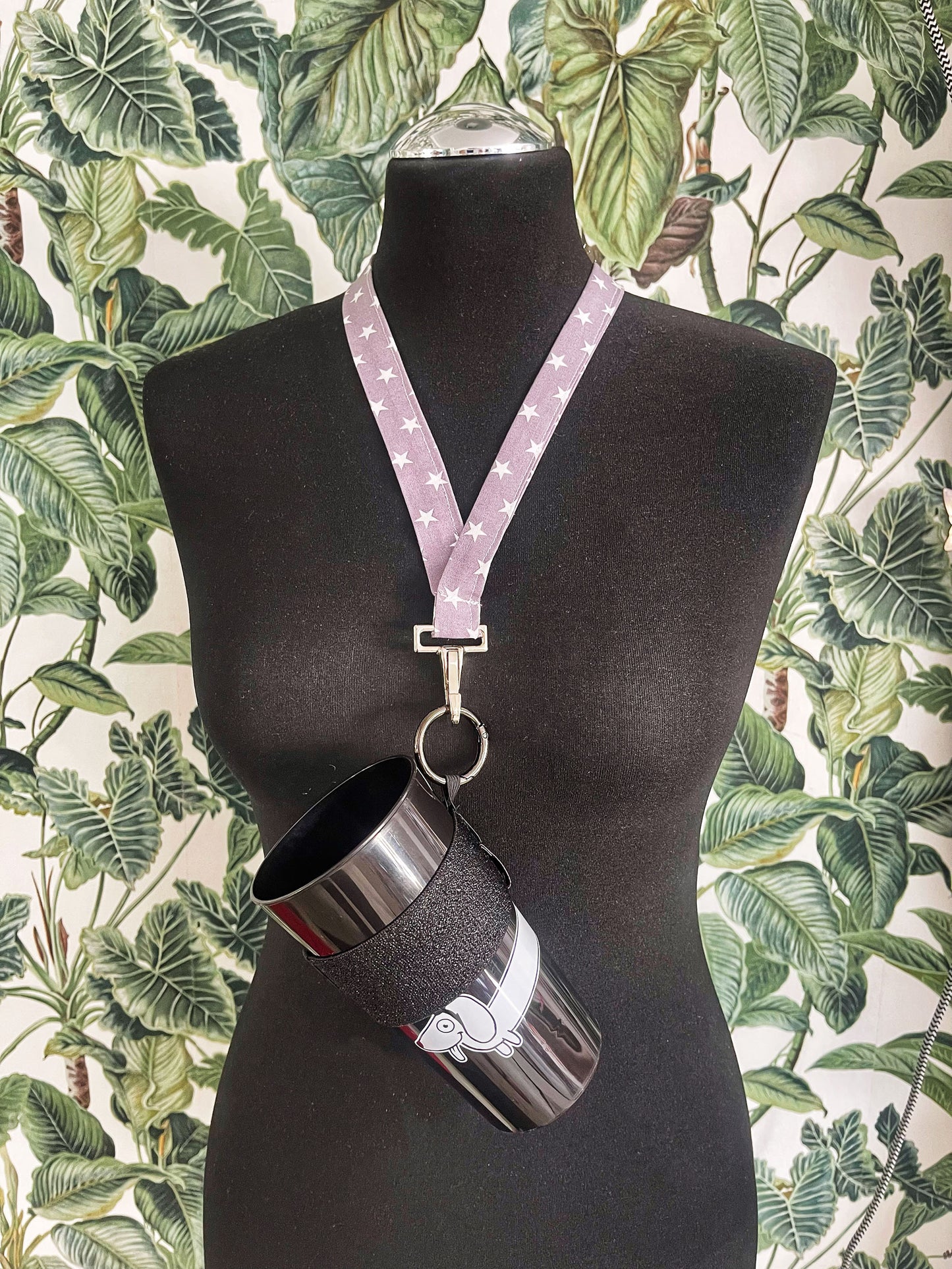 Ein Becher steckt im schwarz glitzernden Becherhalter. Der Karabinerhaken ist an einem Schlüsselband befetigt, das um den Hals einer schwarzen Schneiderpuppe hängt. Die Figur steht vor einer Wand mit grüner Blättertapete.