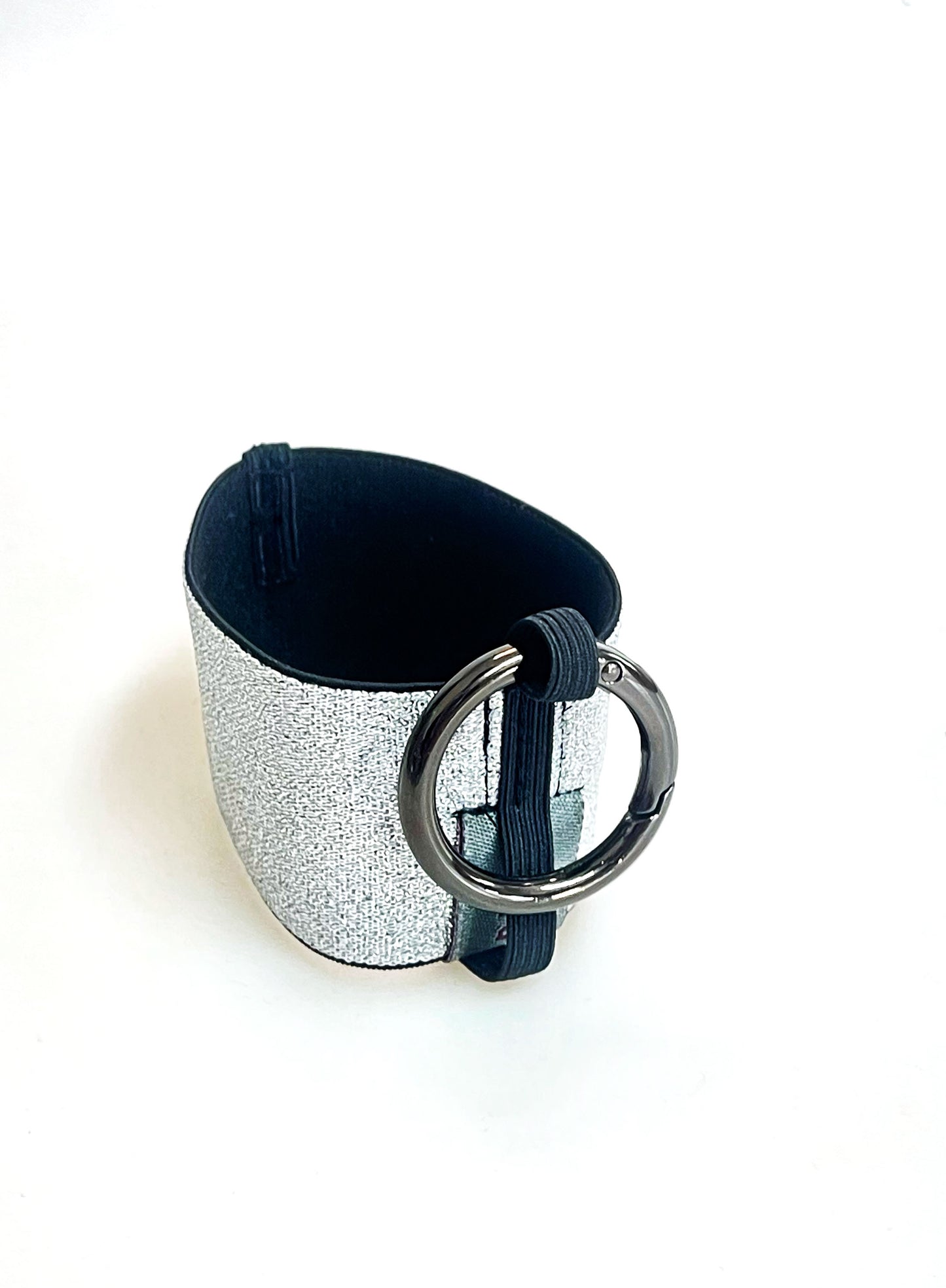 Ein Becherhalter aus einem silber glitzernden, breiten Gummiband, innen schwarz, mit einem runden, gunmetal farbenen Karabinerhaken. Das Gummiband ist zu einem Kreis zusammengenäht. Der Karabiner ist an einem Schmalen Gummiband befestigt, das unten um das breite Gummiband herum geht.