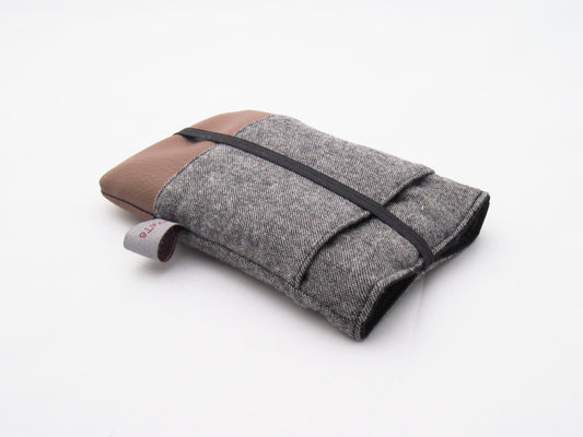 Eine Handytasche aus grauem Tweedstoff oben und braunem Kunstleder unten. Die Tasche hat eine aufgesetzte Tasche aus dem Tweedstoff und ist mit schwarzem Gummiband geschlossen. 
Aufnahme seitlich von oben.