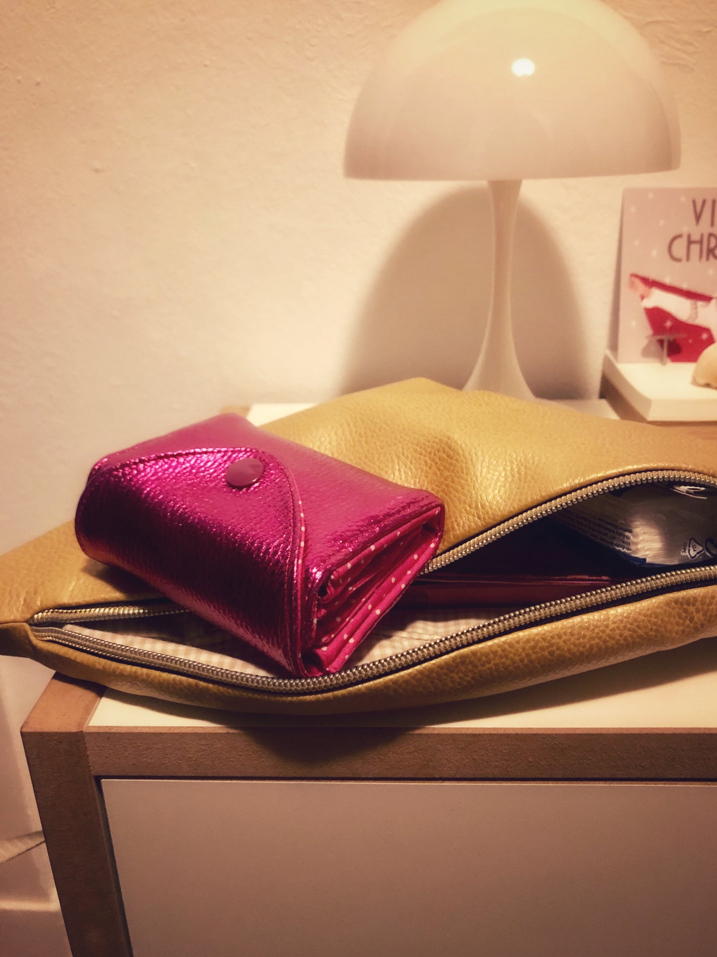 Die pinke Geldbörse auf einer geöffneten, goldenen Bauchtasche liegend. Das Bild ist in einem Zimmer aufgenommen. Tasche und Portemonnaie leigen auf einer Kommode. Im Hintergrud steht eine Lampe.