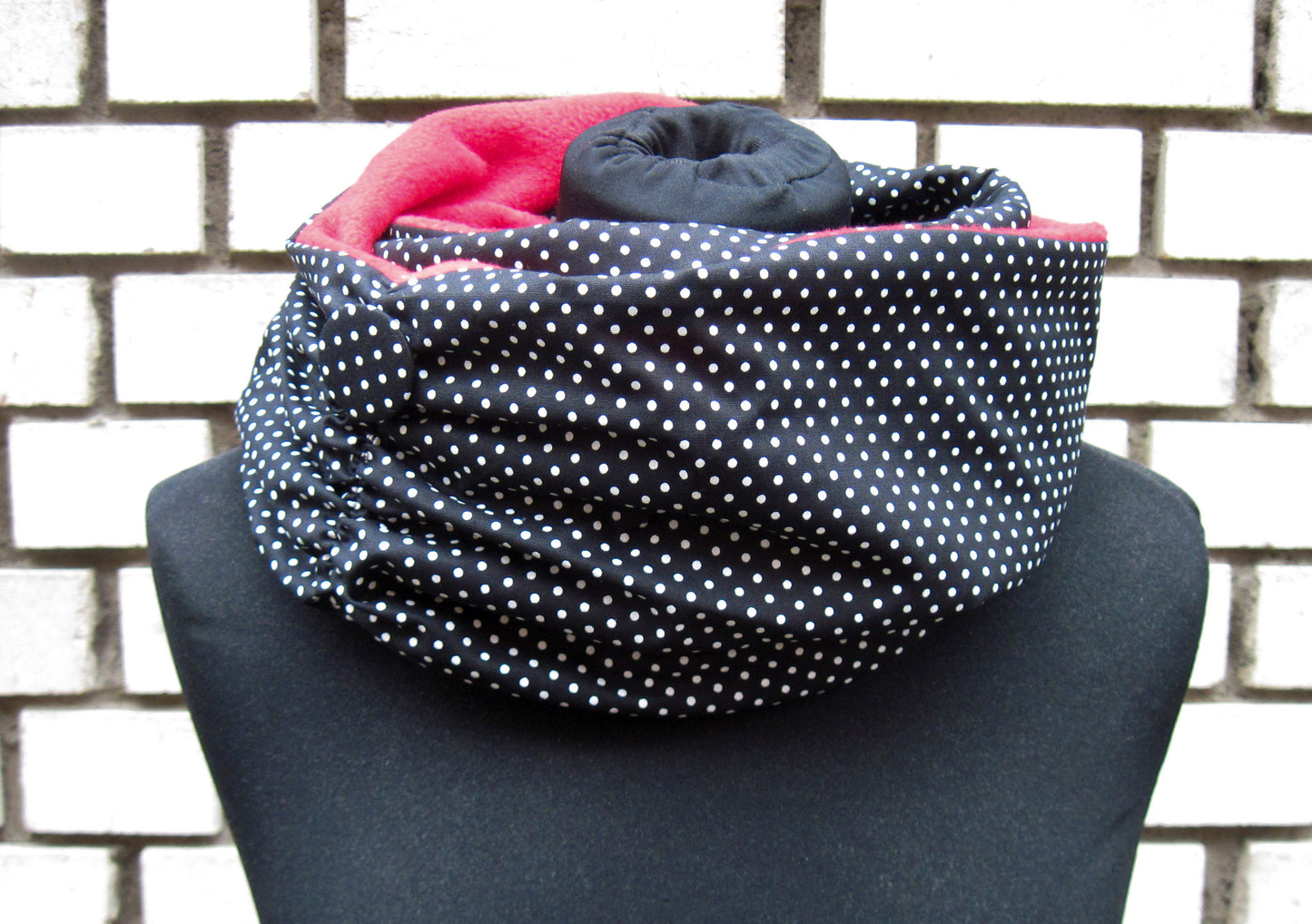 Ein langer Loop-Schal, der zwei Mal um den Hals gewickelt ist. Die äußere Seite des Schals ist aus einem schwarzen Stoff, der mit weißen Pünktchen bedruckt ist. Die innere Seite ist aus rotemPolarfleece. Es schaut oben aus dem Schal hervor. Auf dem Bild links ist der Schal zusammengerafft. An der Stelle hat er auch einen Button aus dem Karostoff. Der Schal ist auf eine schwarzen Schneiderpuppe fotografiert und steht vor einer schwarz-weiß gemauerten Wand.