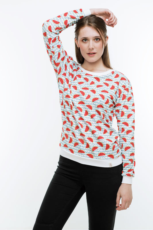 Junge Frau mit langen Haaren in einer engen, schwaren Hose und einem Sweatshirt mit Melonendruck. Es ist weiß und mit roten Wassermelonen und schwarzen Pünktchen auf hellblauen Streifen bedruckt ist. Der Pulli hat weiße Bündchen an Saum, Ärmeln und Halsausschnitt.
Der Halsausschnitt ist rund und etwas größer.