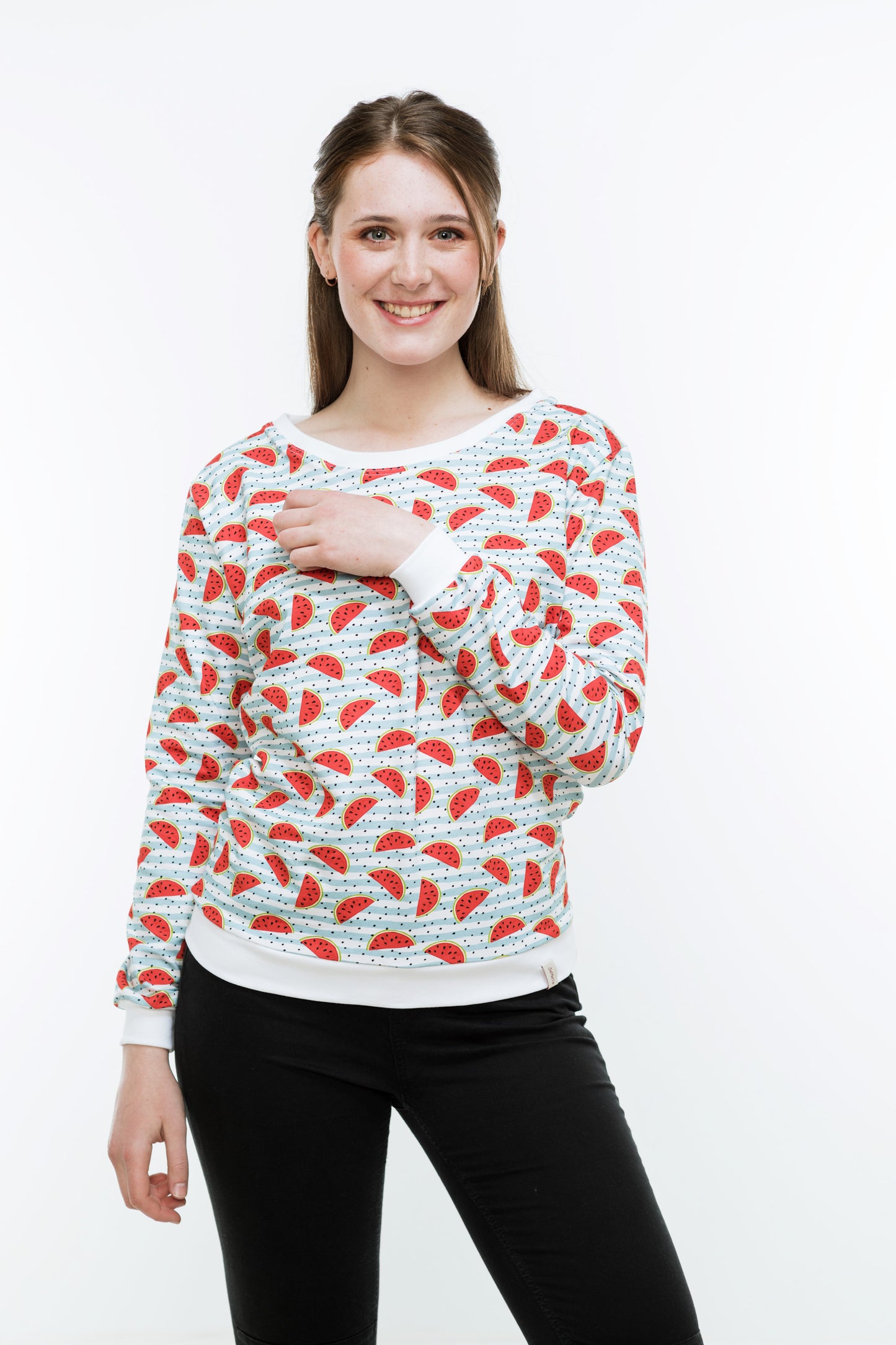 Junge Frau mit langen Haaren in einer engen, schwaren Hose und einem Sweatshirt mit Melonendruck. Es ist weiß und mit roten Wassermelonen und schwarzen Pünktchen auf hellblauen Streifen bedruckt ist. Der Pulli hat weiße Bündchen an Saum, Ärmeln und Halsausschnitt.
Der Halsausschnitt ist rund und etwas größer.