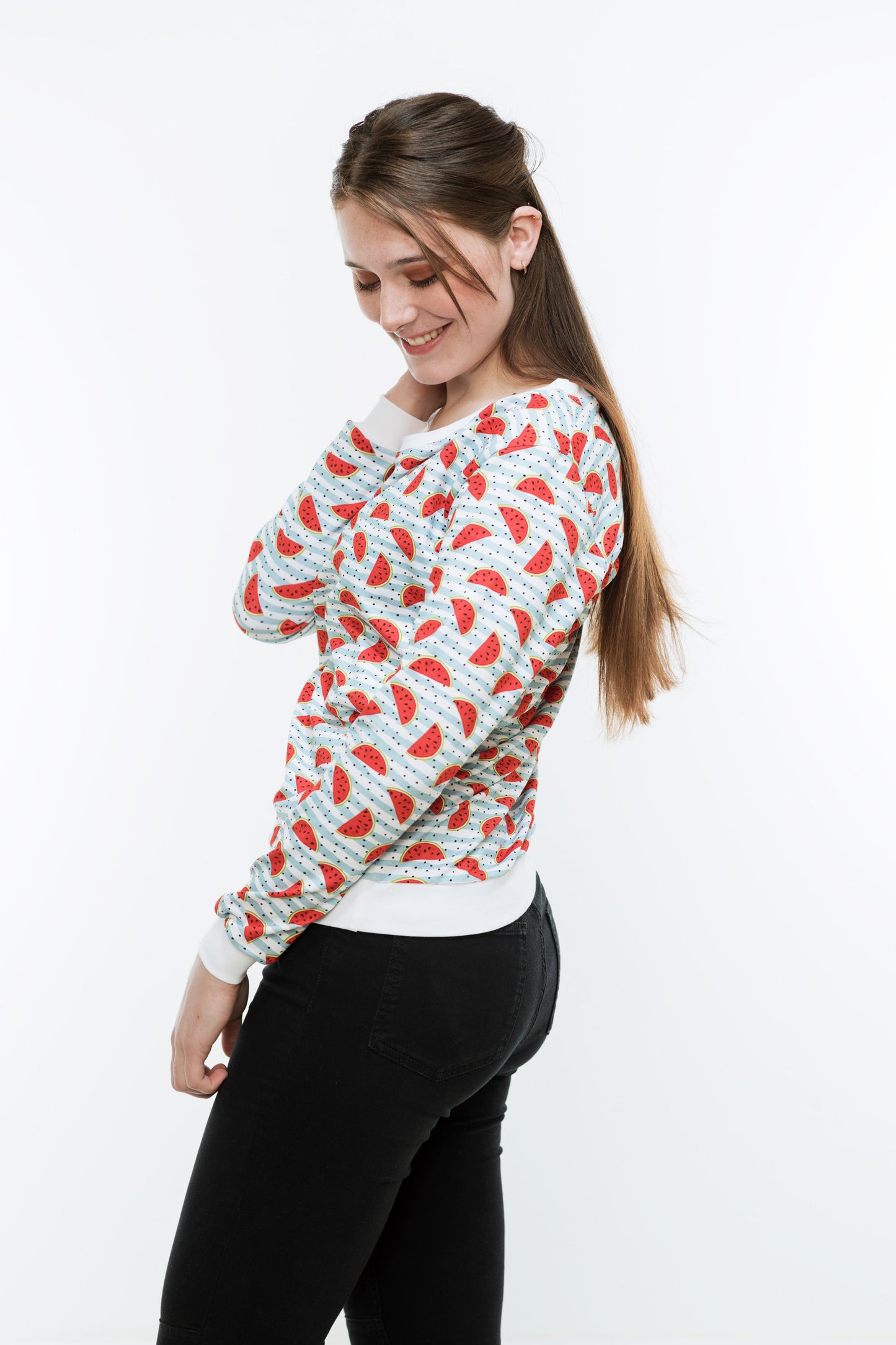 Junge Frau mit langen Haaren in einer engen, schwaren Hose und einem Sweatshirt mit Melonendruck. Es ist weiß und mit roten Wassermelonen und schwarzen Pünktchen auf hellblauen Streifen bedruckt ist. Der Pulli hat weiße Bündchen an Saum, Ärmeln und Halsausschnitt.
Der Halsausschnitt ist rund und etwas größer.
Ansicht von der Seite.