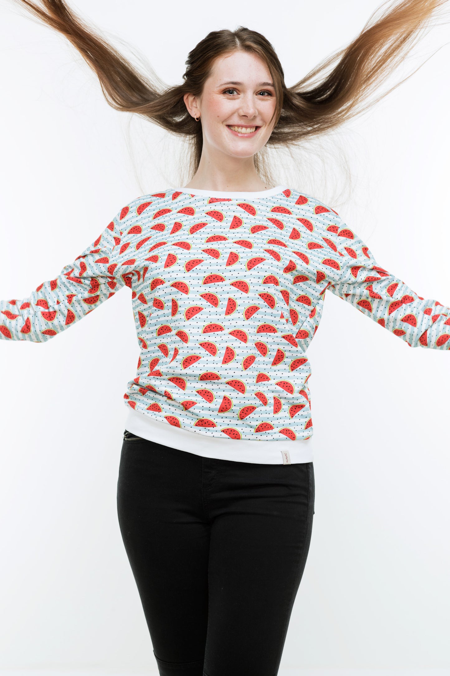 Junge Frau mit langen Haaren in einer engen, schwaren Hose und einem Sweatshirt mit Melonendruck. Es ist weiß und mit roten Wassermelonen und schwarzen Pünktchen auf hellblauen Streifen bedruckt ist. Der Pulli hat weiße Bündchen an Saum, Ärmeln und Halsausschnitt.
Der Halsausschnitt ist rund und etwas größer.
Die Frau wirft die Haare nach oben.