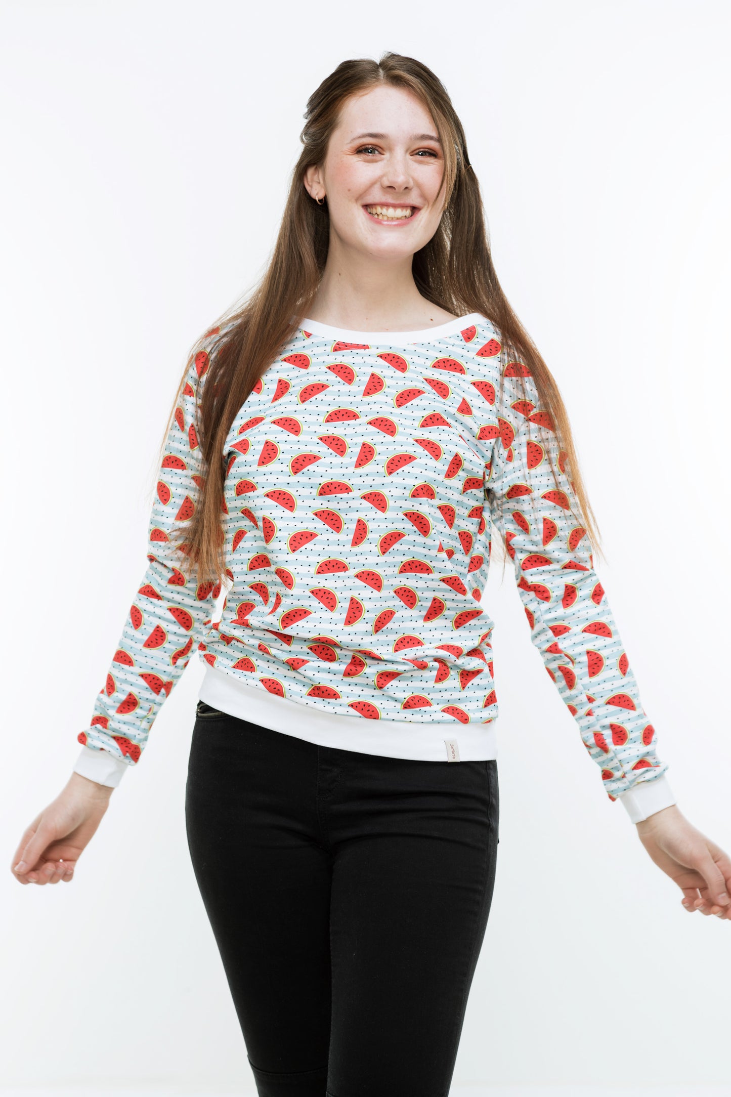Junge Frau mit langen Haaren in einer engen, schwaren Hose und einem Sweatshirt mit Melonendruck. Es ist weiß und mit roten Wassermelonen und schwarzen Pünktchen auf hellblauen Streifen bedruckt ist. Der Pulli hat weiße Bündchen an Saum, Ärmeln und Halsausschnitt.
Der Halsausschnitt ist rund und etwas größer.
Die Frau lacht, die Haare fallen nach vorne.