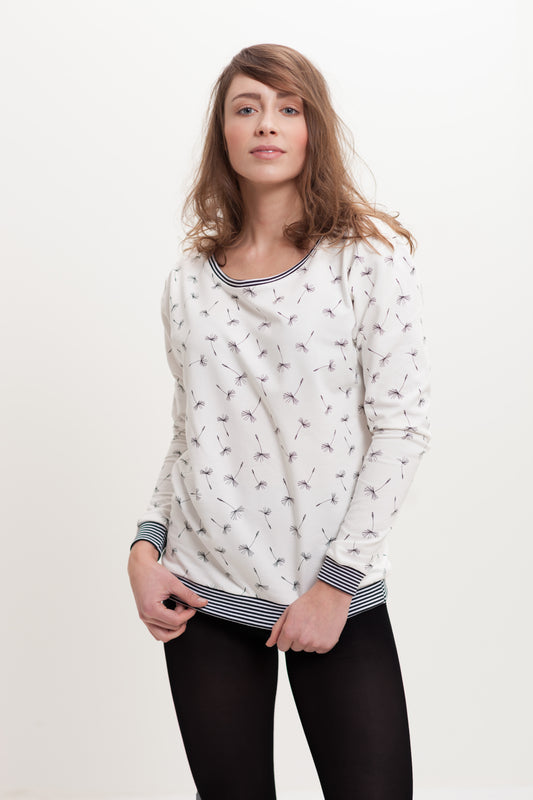 Einen  junge Frau mit langen braunen Haaren. Sie trägt ein ca. hüftlanges weißes Sweatshirt das mit schwarzen Pusteblumendolden bedruckt ist. Das Sweatshirt hat lange Ärmel  und einen weiten Ausschnitt mit schwarz-weißen  Ringelbündchen an Ausschnitt, Ärmeln und Bund. 
Ansicht frontal, leicht seitlich.