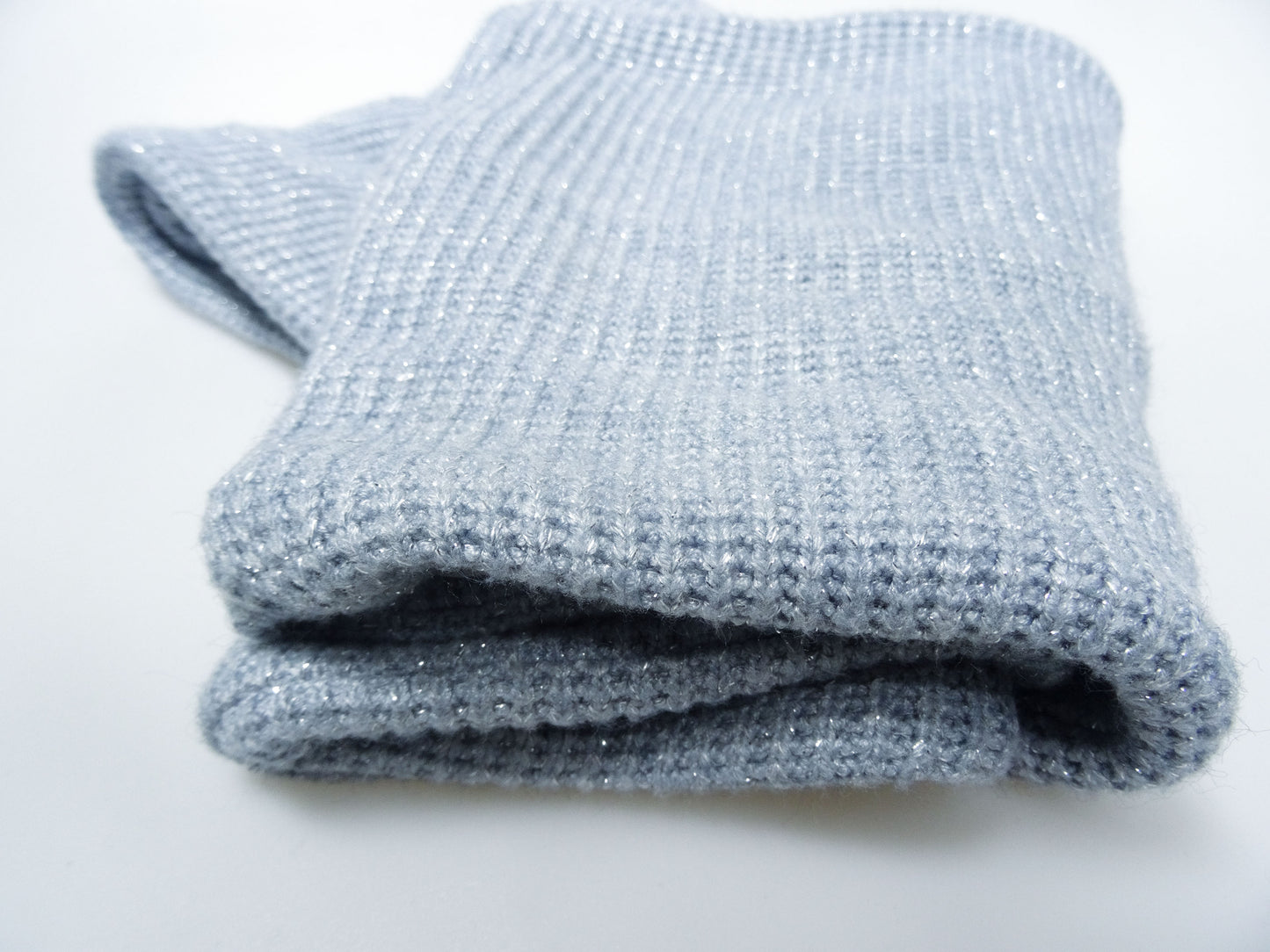 Der hellblaue Loopschal aus Grobstrick mit silbernen Lurxfäden liegt längs gefaltet auf weißem Untergrund. Nahaufnahme von vorne, damit man den Strick gut sieht.