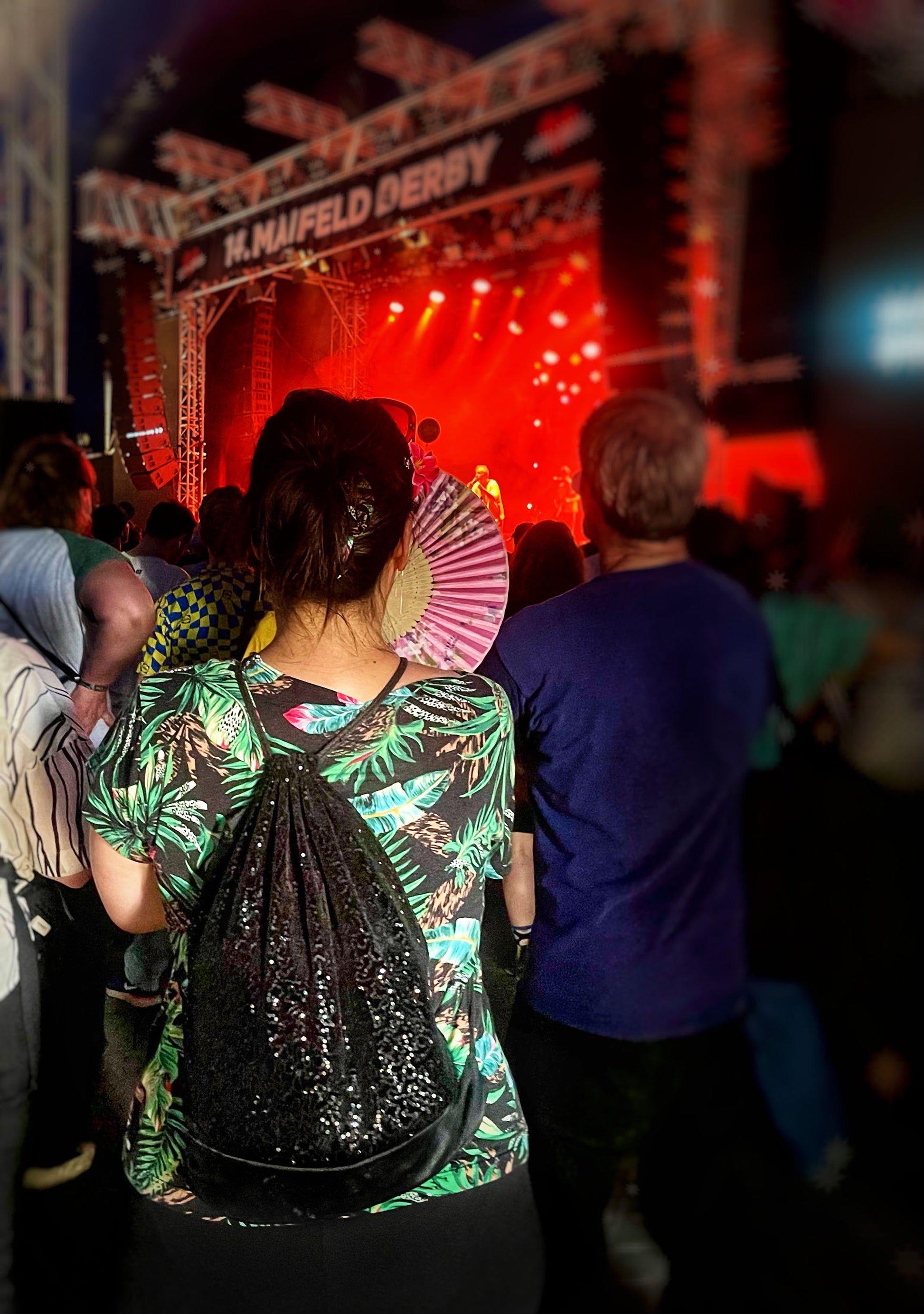 Ene Frau mit dunklen, hochgesteckten Haaren und grün gemustrtem T-Shirt trägt den Turnbeutel mit den Pailletten auf dem Rücken. Sie steht in einer Menschenmenge vor einer Festivabühne und hat einen aufeglappten Fächer in der Hand. Auf der Bühne sind rote Lichter zu sehen und der Schiftzug "14. Maifeld Derby". Man sieht die Frau und die Mescnenmenge von hinten.