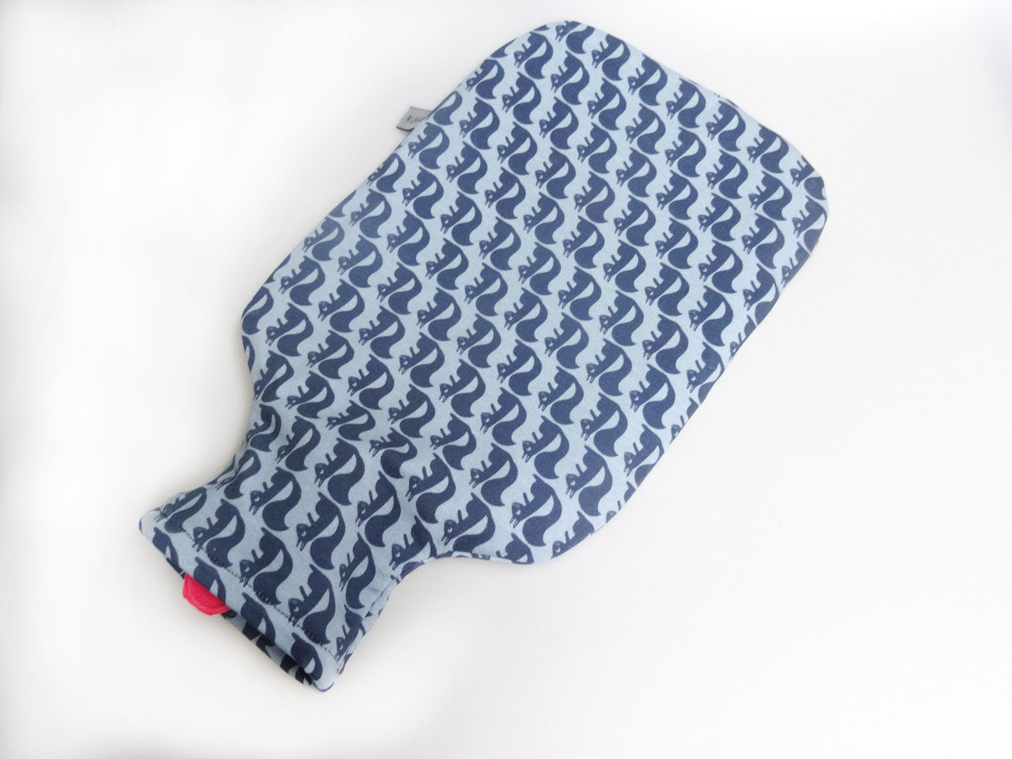 eine Wärmflaschen mit einer Wärmflaschenhülle aus hellblauem Jersey, die mit dunkelblauen, gezeichneten Eichhörnchen bedruckt ist. Die Eichhörnchen schauen alle in die selbe Richtung. Das Muter wirkt grafisch. An der oberen Öffnung ist die rote Gummiwärmflaschen zu sehen.