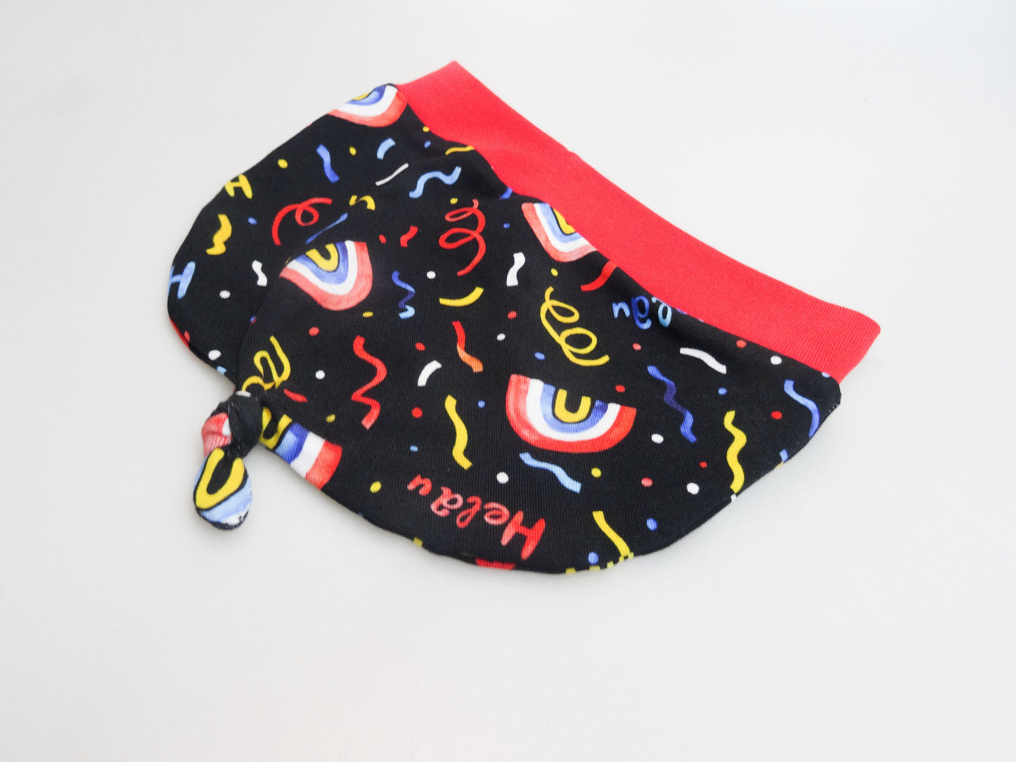 Zipfelmütze für Babys und kleine Kinder schwarz mit buntem Schriftzug Helau, Konfetti, Luftschlangen und Regenbögen. Ansicht liegend seitlich Zipfel unten.