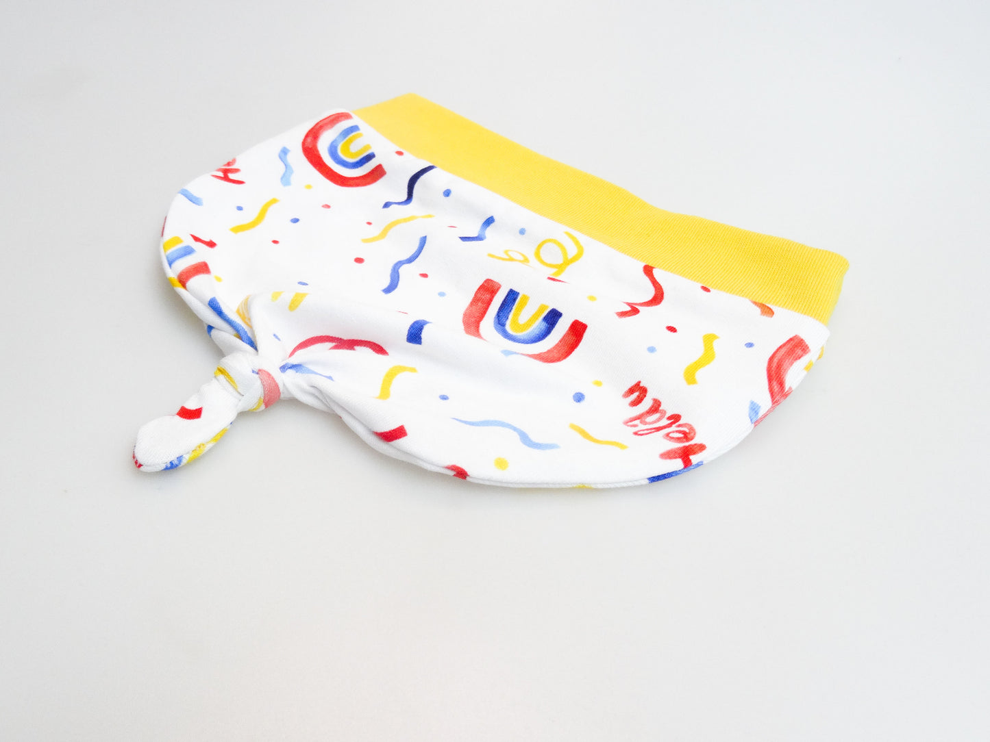Zipfelmütze für Babys und kleine Kinder weiß mit buntem Schriftzug Helau, Konfetti, Luftschlangen und Regenbögen. Ansicht liegend seitlich Zipfel unten.