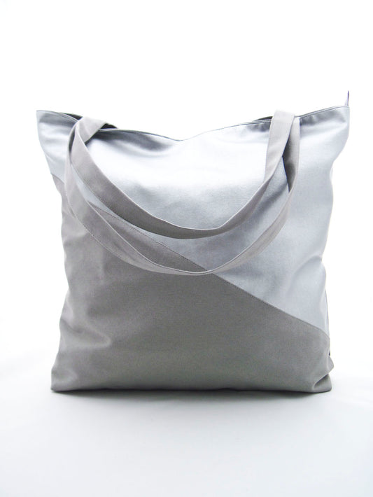 Ein Shopper aus Kunstleder und Canvas vor einem weißen Hintergrund. Die Tasche ist diagonal geteilt. Der untere Teil und die Träger sind aus grauem Canvas. Der oberer Teil ist aus silbernem Kunstleder. Die Tasche ist rechteckig und höher, als breit.