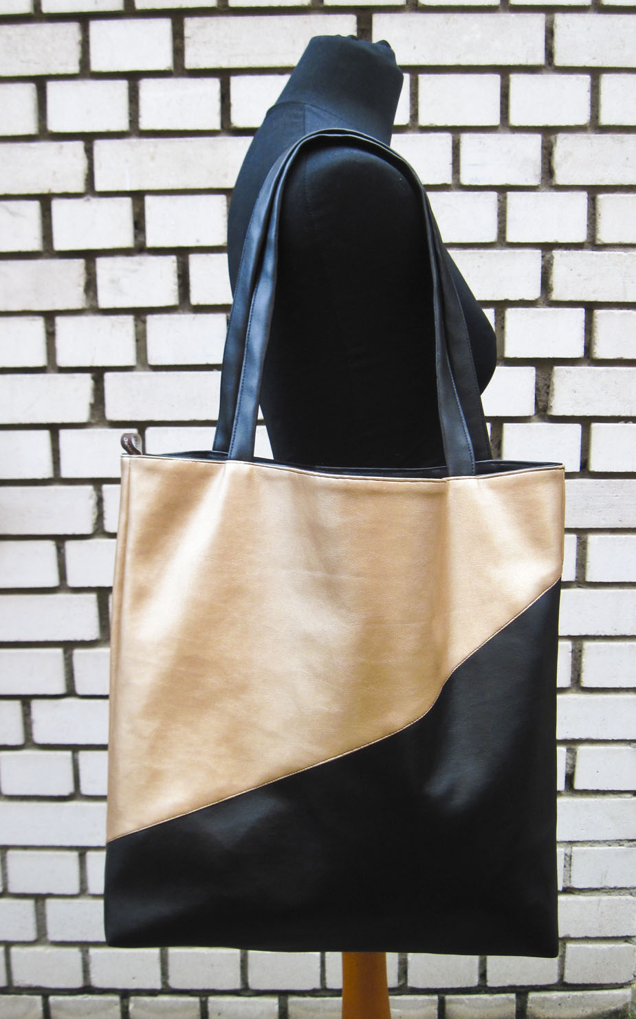 Der diagonal geteilte, schwarz-goldene Shopper auf der Schulter einer schwarzen Schneiderpuppe vor einer schwarz-weiß gemauerten Wand.