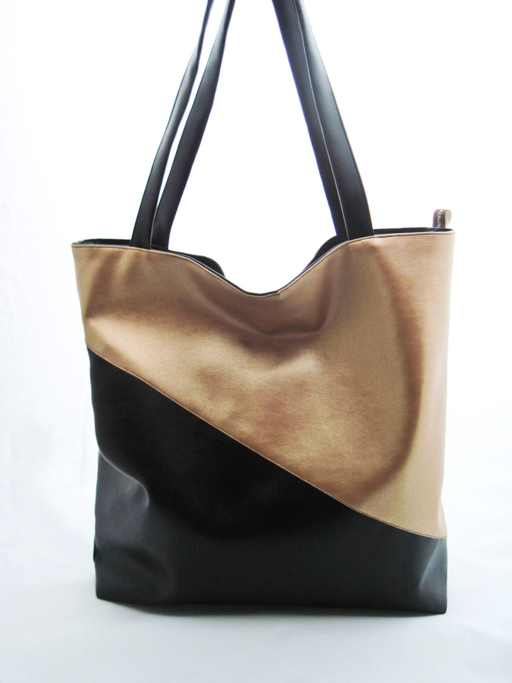 Ein Shopper aus Kunstleder vor einem weißen Hintergrund. Die Tasche ist diagonal geteilt. Der untere Teil und die Träger sind schwarz. Der oberer Teil ist gold. Die Tasche ist rechteckig und höher, als breit.