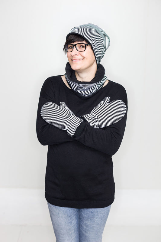 Eine Frau mit Brille und dunkler Ponyfrisur. Sie trägt eine schwarz-weiß geringelte Mütze und einen kurzen Loopschal, der auf einer Seite schwarz-weiß geringelt ist und auf der anderen Seite aus schwarzen Fleece. Passend dazu trägt sie auch Fäustlinge aus dem schwarz-weiß geringekten Stoff. 
Außerdem hat sie inen schwaqrzen Pullover und hellblaue Jeans an.