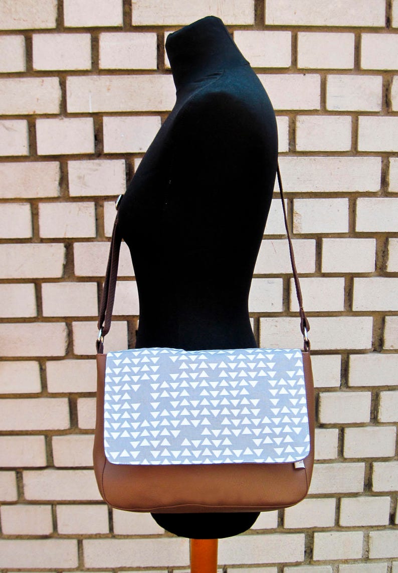 Die braunem Messenger Tasche aus Kunstleder mit einem Deckel aus grauem Stoff mit weißen Dreiecken seitlich auf einer Schneiderpuppoe hängend, vor einer Backsteinwand. Die Tasche hat einen braunen Tragegurt mit silbernen D-Ringen und einem silbernen Versteller.