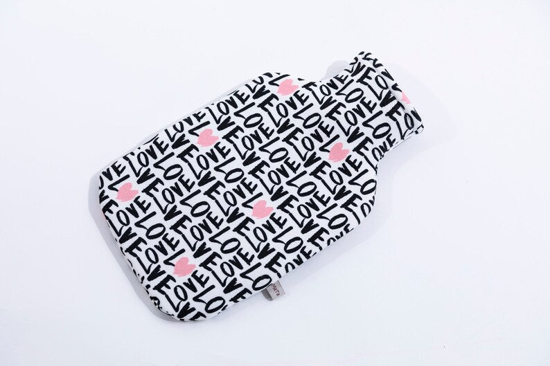 Eine liegende, weiße Wärmflaschenhülle mit schwarzem "Love" Schriftzug und rosa Herzen. Ansicht von oben, seitlich nach rechts gelegt.