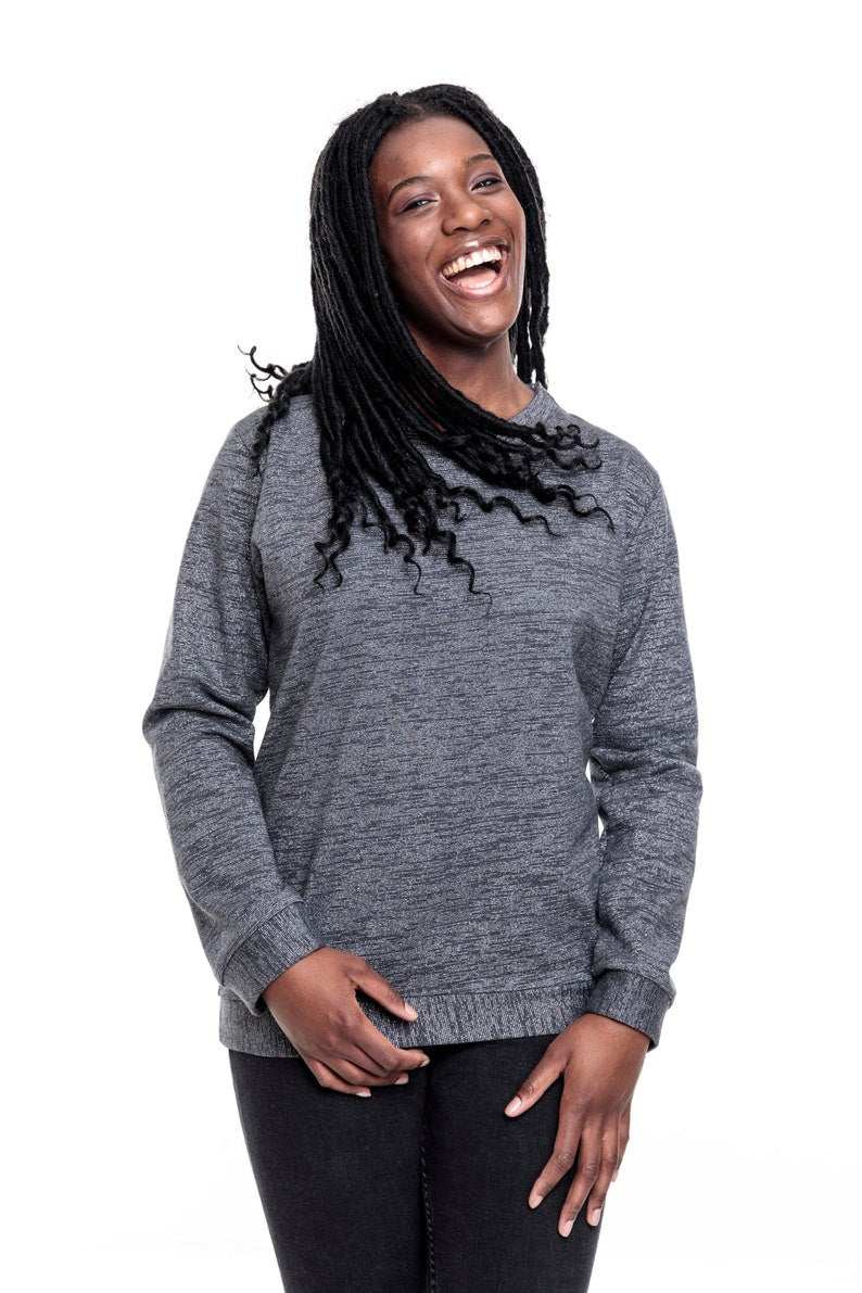 Eine junge Frau mit langen Braids. Sie trägt ein graues, meliertes Sweatshirt mit silbernen Lurexfäden. Das Sweatshirt ist ca. hüftlang, hat lange Ärmel und einen Runden Halsausschnitt. Als Abschluss an den Ärmeln, dem Bund und dem Hals hat der Pulli Bündchen aus dem gleich Stoff. 
Ansicht frontal, in der Bewegung.