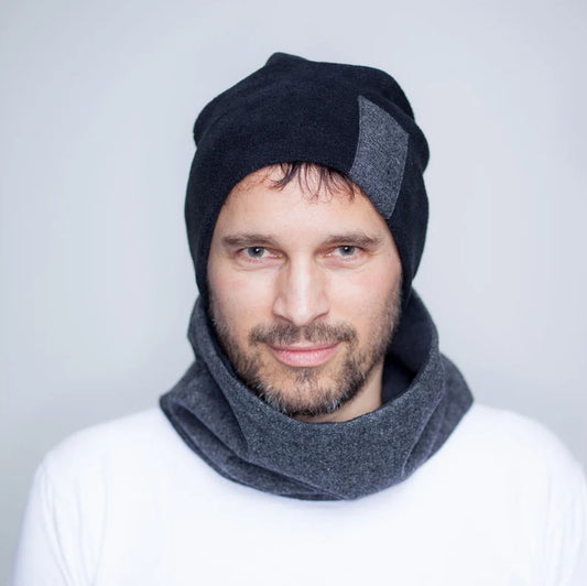 Ein Mann mit dunklen Haaren und Bart. Er trägt eine Beaniemütze aus schwarzem Fleece mit einem Rechteck aus grauem Wollstoff auf der linken Seite. Passend dazu trägt er einen kurzen Loopschal, der auf der einen Seite aus dem grauen Wollstoff und auf der anderen Seite aus schwarzem Fleece ist. Der graue Wollstoff hat eine klassiche Pfeffer und Salz Optik.