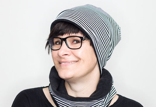 Close Up einer dunkelhaarigen Frau mit Ponyfrisur und Brille. Sie trägt eine schwarz-weiß geringelte, lange Neanie Mütze. Am Rand der Mütze ist das schwarze Innenteil aus Polarfleece zu sehen. Dazu trägt sie einen passenden, kurzen geringelten Loop Schal mit Fleece Futter .