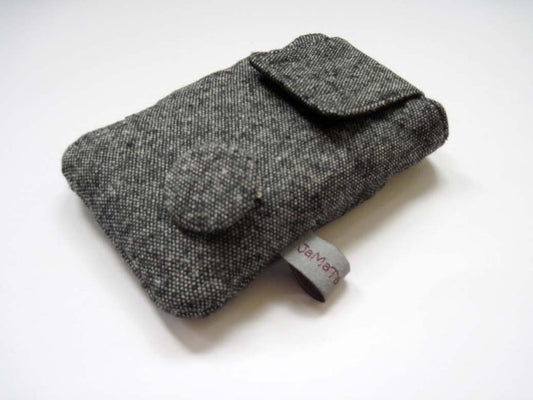 Eine Handytasche aus grauem Tweed in Pfeffer und Salz Optik. Die Tasche hat eineKlappe zum Verschließen und unten rechte einen Button aus dem Tweedstoff. Aufnahme seitlich von unten.