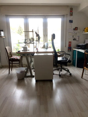 Aufnahme eines Zimmers mit hellem Laminatboden. Vor dem Fenster stehen zwei Nähmaschinen jeweils auf einem Tisch. Vor den Nähtischen stehen Stühle, an der rechten Wand ist ein weiterer Tisch und Stuhl zu erkennen und ein kleiner Schubladenschrank. An der Wand neben dem fenster hängen Postkarten. Auf dem Tisch an der Wand steht ein Drucker.