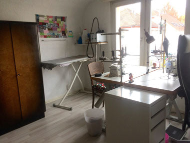 Die linke Seite des Raums. Man sieht die Nähmaschinen avor dem Fenster und die Stühle davor. In der Ecke neben dem Fenster steht ein Bügelbrett und auf einem Regal darüber das Bügeleisen. Über dem Bügelbrett hängt ein Bild. Neben dem Bübeglebrett steht ein dunkelbrauner Schrank.