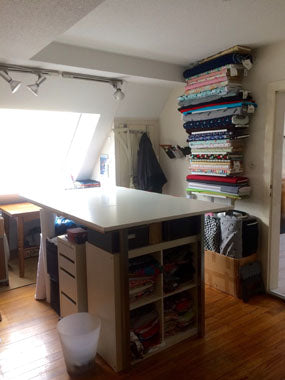 Ein anderes Zimmer mit Dielenboden. In der Mitte steht ein großer Zuschneidetisch mit Regalen darunter. An der Wand ist ein Hängereagl mit Stoffen darauf. Unter dem Regal steht eine Kiste mit Stoffen darin. Links ist ein großesw dachfenster mit einer Schiene darüber, in der drei Scheinwerfer hängen.
