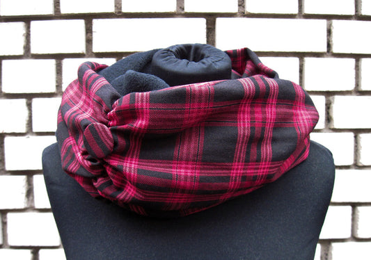 Ein langer Loop-Schal, der zwei Mal um den Hals gewickelt ist. Die äußere Seite des Schals ist aus einem schwarz-pinken Schottenkaro. Die innere Seite ist aus schwarzem Polarfleece. Es schaut oben aus dem Schal hervor. Auf dem Bild links ist der Schal zusammengerafft. An der Stelle hat er auch einen Button aus dem Karostoff. Der Schal ist auf eine schwarzen Schneiderpuppe fotografiert und steht vor einer schwarz-weiß gemauerten Wand.