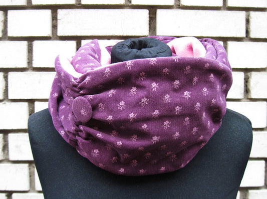 Ein langer Loop-Schal, der zwei Mal um den Hals gewickelt ist. Die äußere Seite des Schals ist aus lila Feincord, der mit rosa Blümchen bedruckt ist. Die innere Seite ist aus rosa Polarfleece. Es schaut oben aus dem Schal hervor. Auf dem Bild links ist der Schal zusammengerafft. An der Stelle hat er auch einen Button aus dem Cordstoff. Der Schal ist auf eine schwarzen Schneiderpuppe fotografiert und steht vor einer schwarz-weiß gemauerten Wand.
