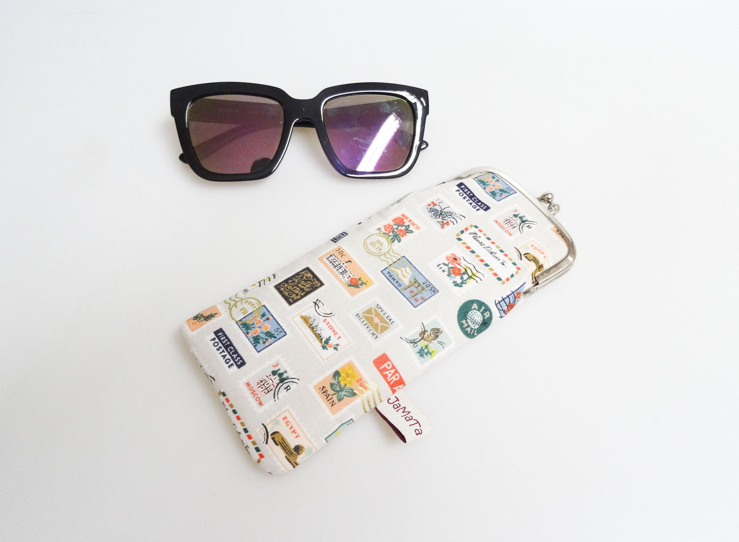 Ein Brillenetui mit retro Briefmarkenmotiv und silbernem Clipverschluss. Darüber eine Sonnenbrille mit schwarzem Rahmen.