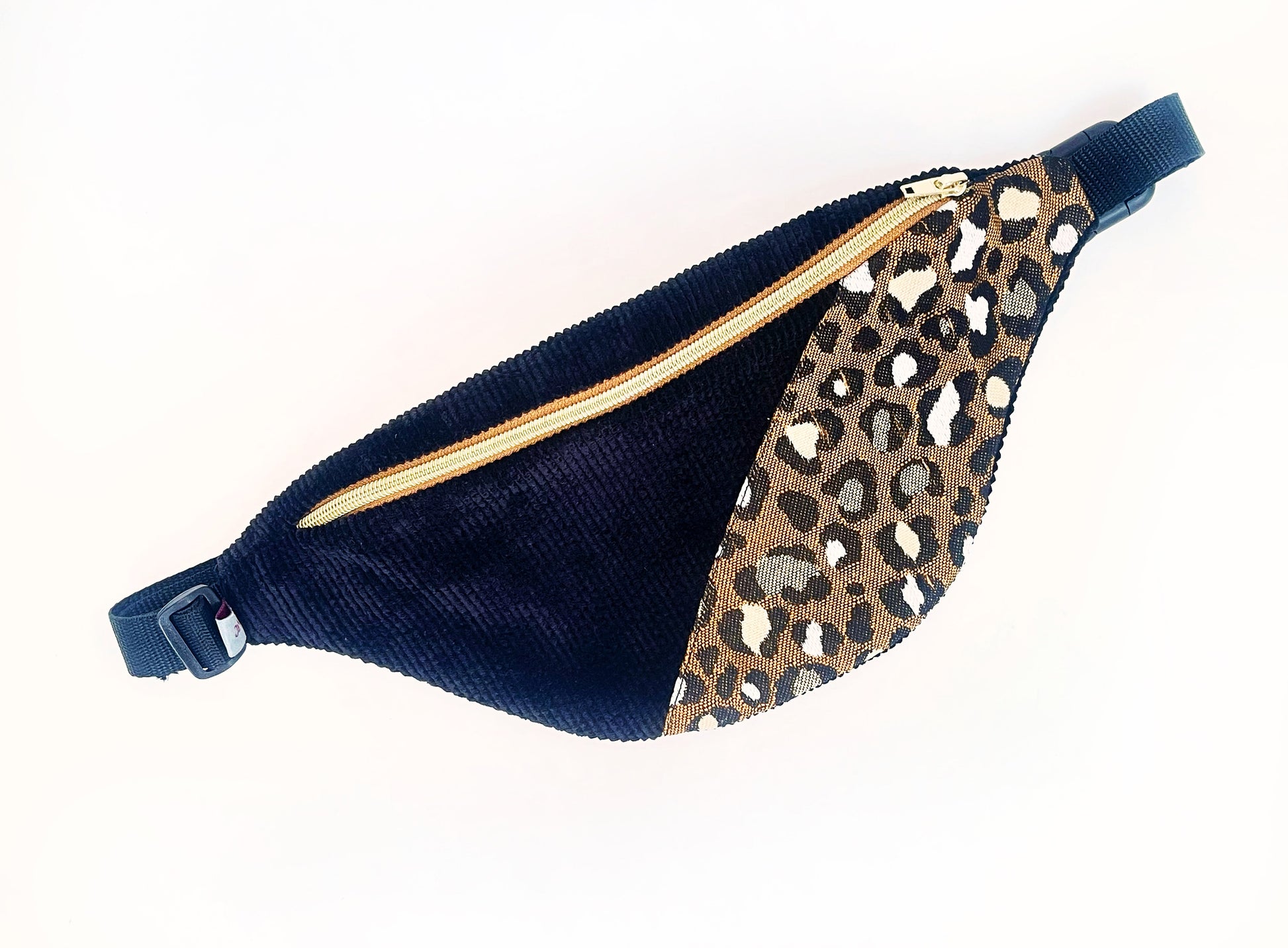 Eine Bauchtasche in abgerundeter Form. Das Vorderteil ist geteilt. Der linke Teil ist aus schwarzem Breitcord und der rechte aus einem Jacquard Canvas im Leopradenprint. Die Tasche wird mit einem goldenen Reißverschluss auf braunem Band geschlossen. Der Deckel der Tasche ist aus dem schwarzen Breitcord. Sie hat schwarzes Gurtband, eine Steckschnalle und einen Versteller aus schwarzem Kunststoff. 
Ansicht von oben, schräg. 
