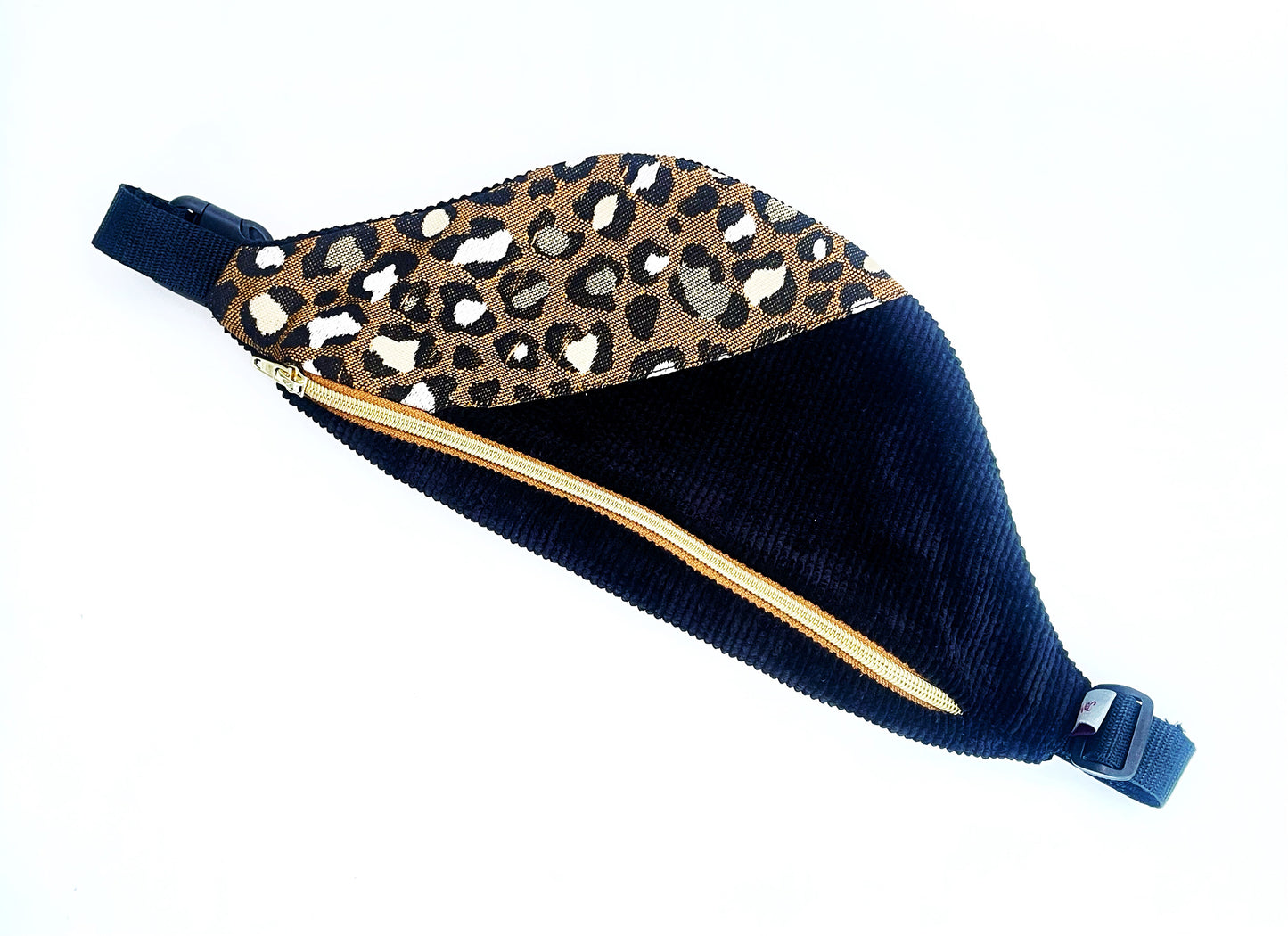 Eine Bauchtasche in abgerundeter Form. Das Vorderteil ist geteilt. Der linke Teil ist aus schwarzem Breitcord und der rechte aus einem Jacquard Canvas im Leopradenprint. Die Tasche wird mit einem goldenen Reißverschluss auf braunem Band geschlossen. Der Deckel der Tasche ist aus dem schwarzen Breitcord. Sie hat schwarzes Gurtband, eine Steckschnalle und einen Versteller aus schwarzem Kunststoff. 
Ansicht von oben, schräg. De Tasche ist gedreht, so dass der Deckel unten im Bild ist.
