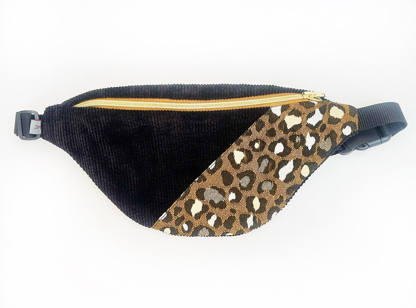 Eine Bauchtasche in abgerundeter Form. Das Vorderteil ist geteilt. Der linke Teil ist aus schwarzem Breitcord und der rechte aus einem Jacquard Canvas im Leopradenprint. Die Tasche wird mit einem goldenen Reißverschluss auf braunem Band geschlossen. Der Deckel der Tasche ist aus dem schwarzen Breitcord. Sie hat schwarzes Gurtband, eine Steckschnalle und einen Versteller aus schwarzem Kunststoff. 
Ansicht von oben, gerade. 