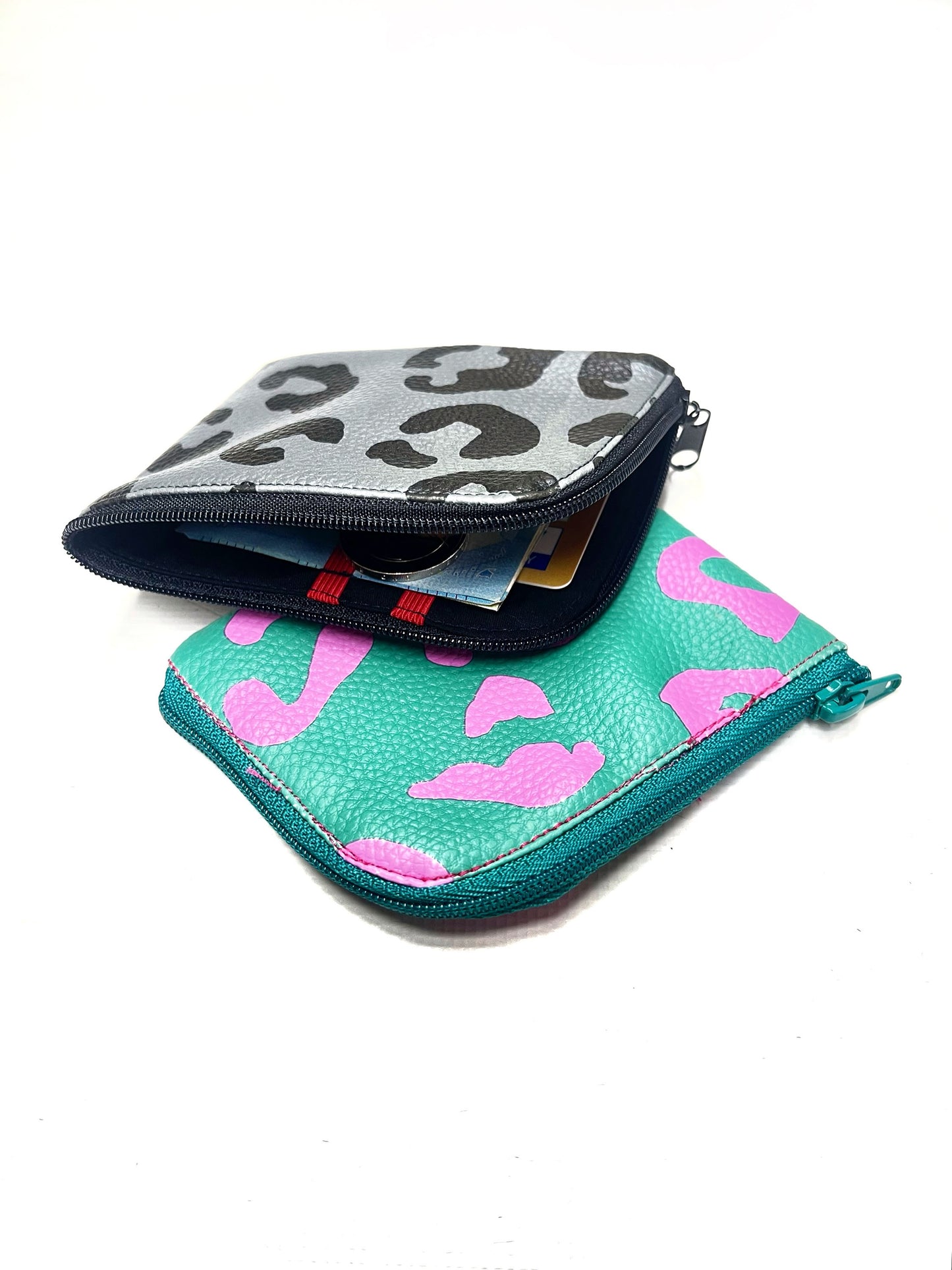 Die graue und die grüne Geldbörse liegen übereinander. Die grüne liegt unten, mit der abgerundeten Ecke nach vorne. Die graue liegt halb darüber und ist geöffnet. Dadurch kann man Geld und eine Karte im Inneren erkennen und auch die roten Gummibänder. 